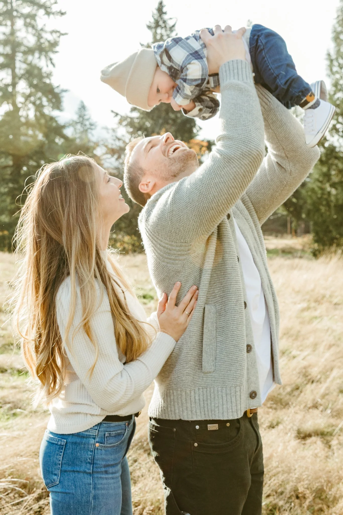 A family outdoors, a man lifting a young boy into the air while a woman smiles and touches his back, in a grassy area with trees, sunny weather.