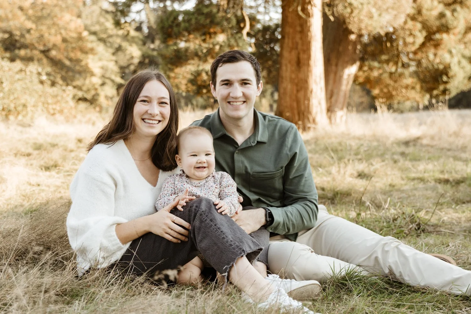 A happy family of three sitting on grass in a park with trees in the background during autumn. The mother has long brown hair, wearing a white sweater. The father has short dark hair, wearing a green shirt. The baby girl is smiling, sitting between h