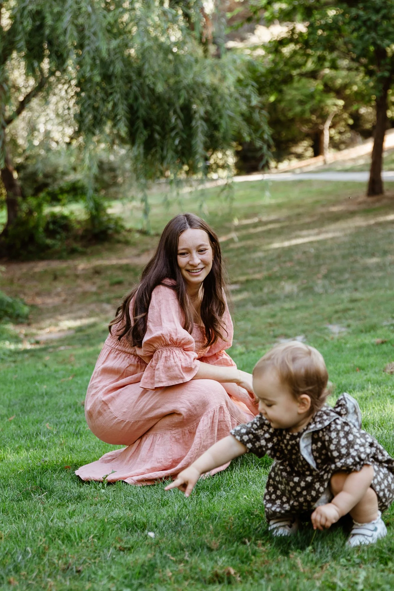A young woman with long brown hair wearing a pink dress squats on grass, smiling, as she watches a toddler with short blonde hair wearing a floral dress and white shoes crawling and pointing at something on the ground in a park or garden with trees a