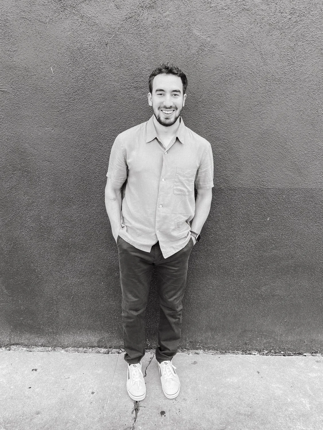 A young man with a beard and short hair stands against a textured wall, smiling and looking at the camera, with his hands in his pants pockets, wearing a short-sleeved button-up shirt, dark pants, and sneakers.