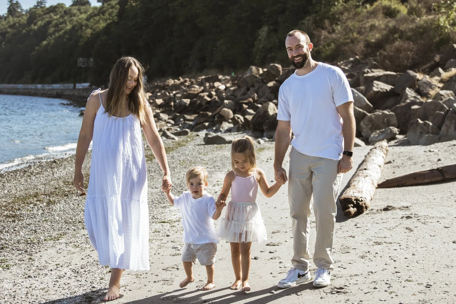 A family of four walking hand in hand on a beach, with rocky shoreline and green hillside in the background.