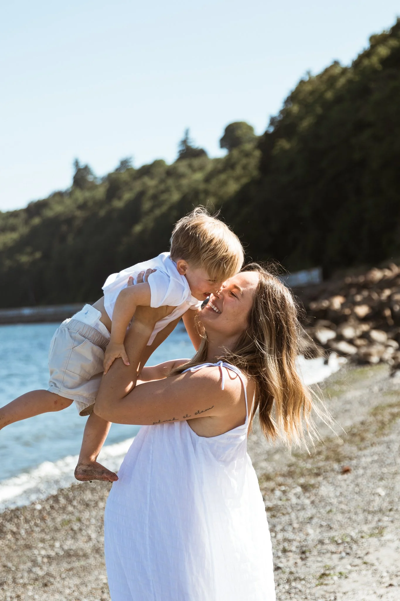 A woman holding a young boy on a beach with trees in the background. They are smiling and appear to be playing or enjoying each other's company.