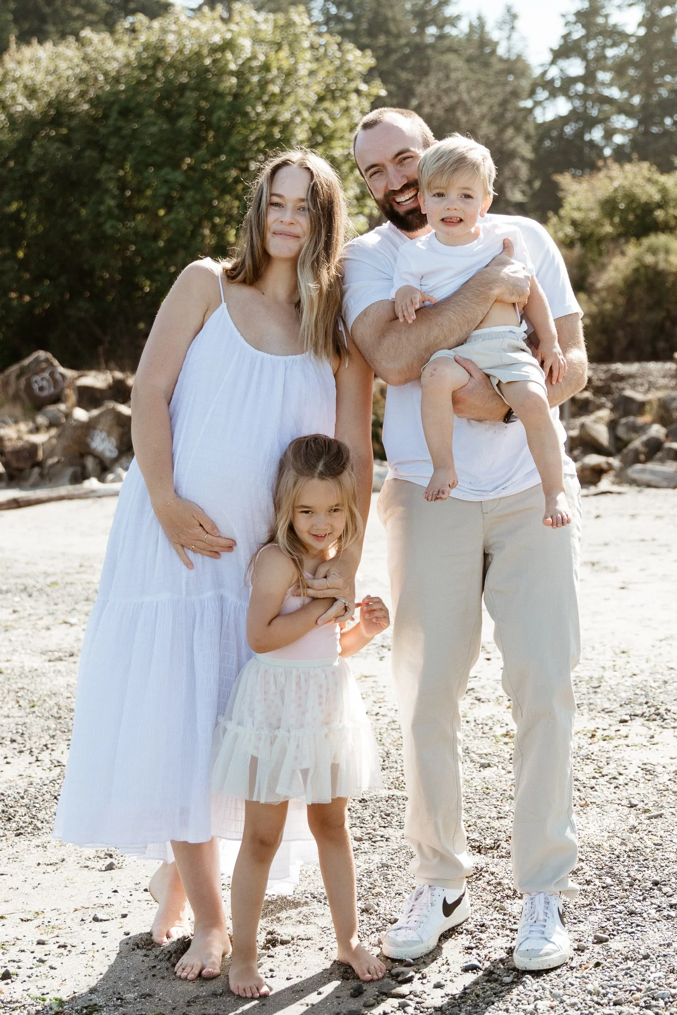 A family of five standing outdoors on a beach, smiling for a photo. The mother has long light brown hair and is wearing a white sundress. The father has a beard and is wearing a white T-shirt and light-colored pants, holding a young boy with blonde h