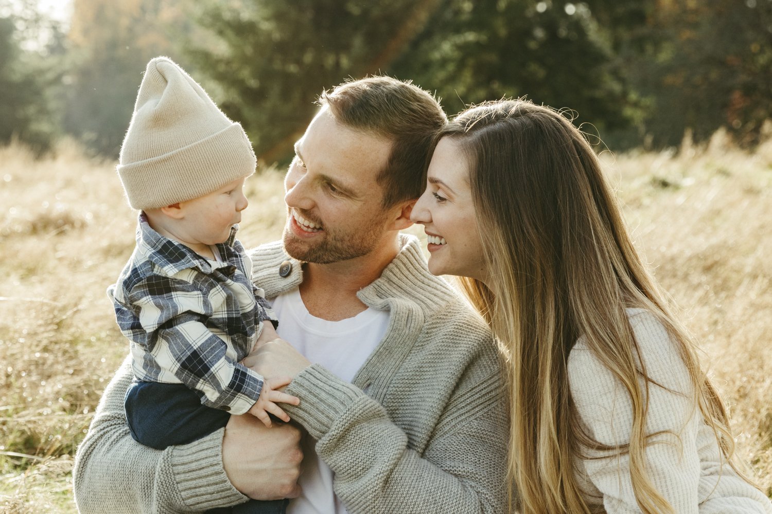 A family of three outdoors on a sunny day, with the father holding a young boy wearing a beige beanie and plaid shirt, and the mother smiling at them in a light-colored sweater, all enjoying a moment together.
