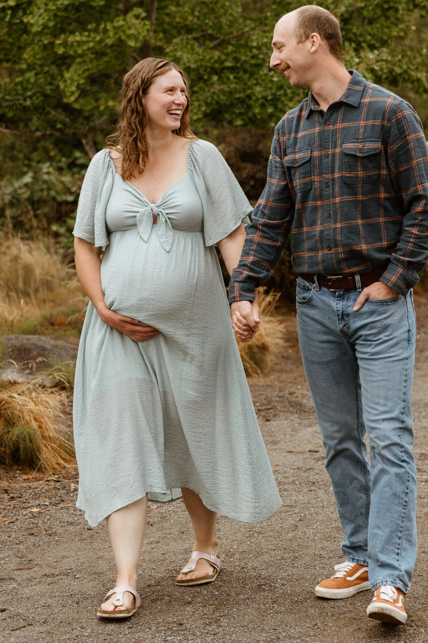 A pregnant woman and a man hold hands and walk outdoors on a dirt path, smiling at each other.