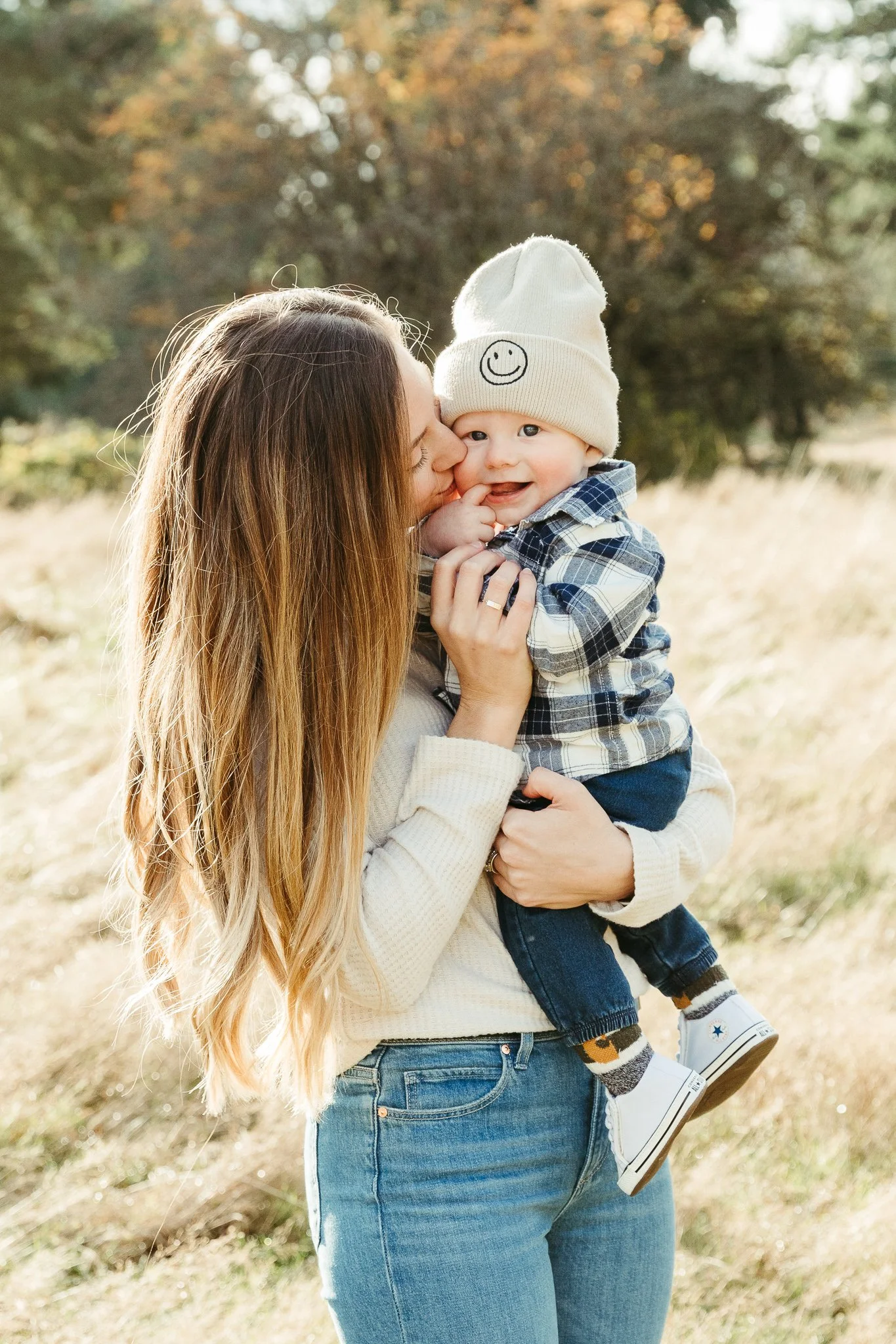 A woman holding a smiling baby boy outdoors during autumn, with trees in the background. The baby is wearing a white beanie with a smiley face and a plaid shirt.