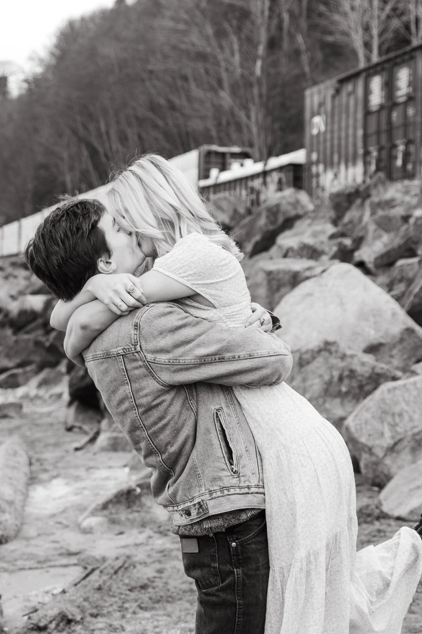 A black and white photo of a couple kissing on a rocky beach, with the woman being lifted by the man.
