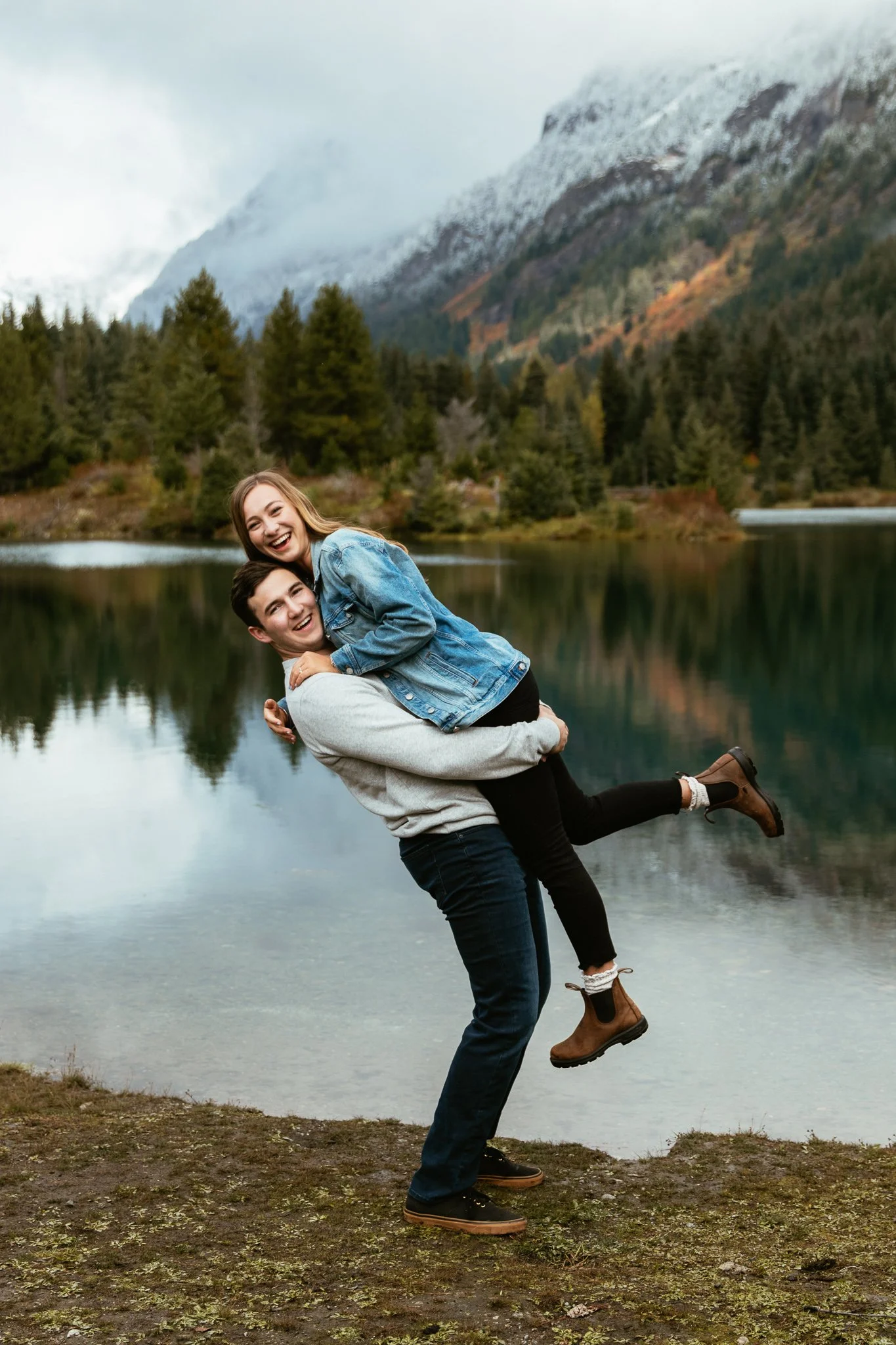 A couple is outdoors near a lake, with the woman on the man's back, both smiling and enjoying the moment. The background shows trees and mountains.