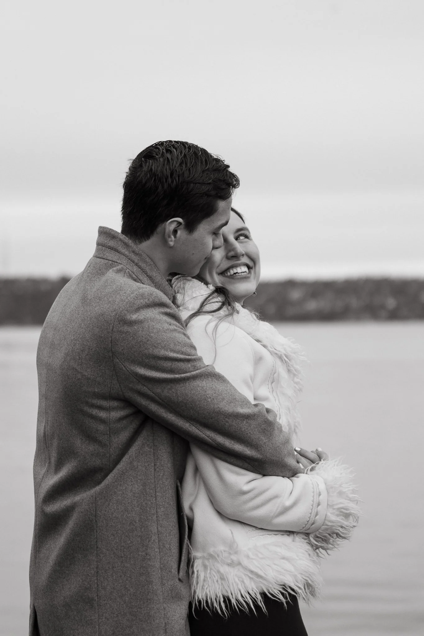 A black and white photo of a couple embracing by a body of water, with the man kissing the woman's forehead as she smiles.
