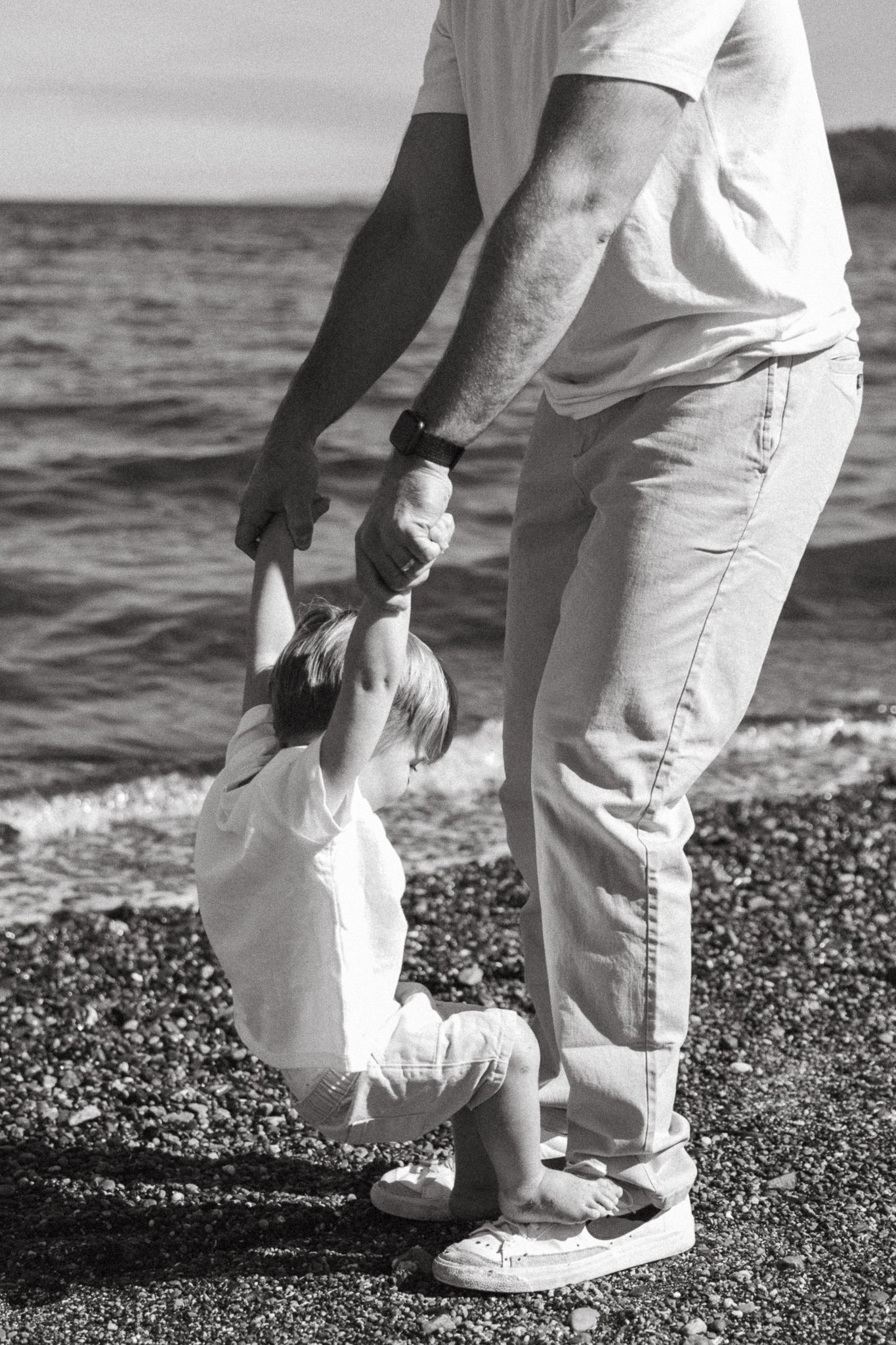 A black and white photo of a child holding hands with an adult, standing on a pebbled beach by the water.