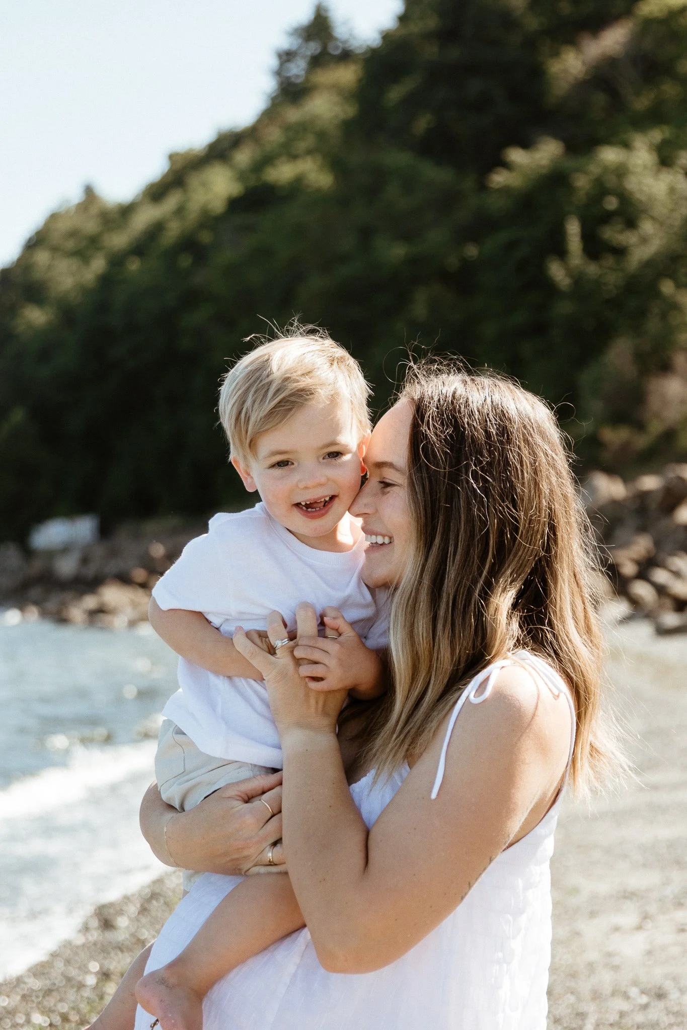 A woman and young boy smiling and playing at the beach, with rocky shoreline and green trees in the background.