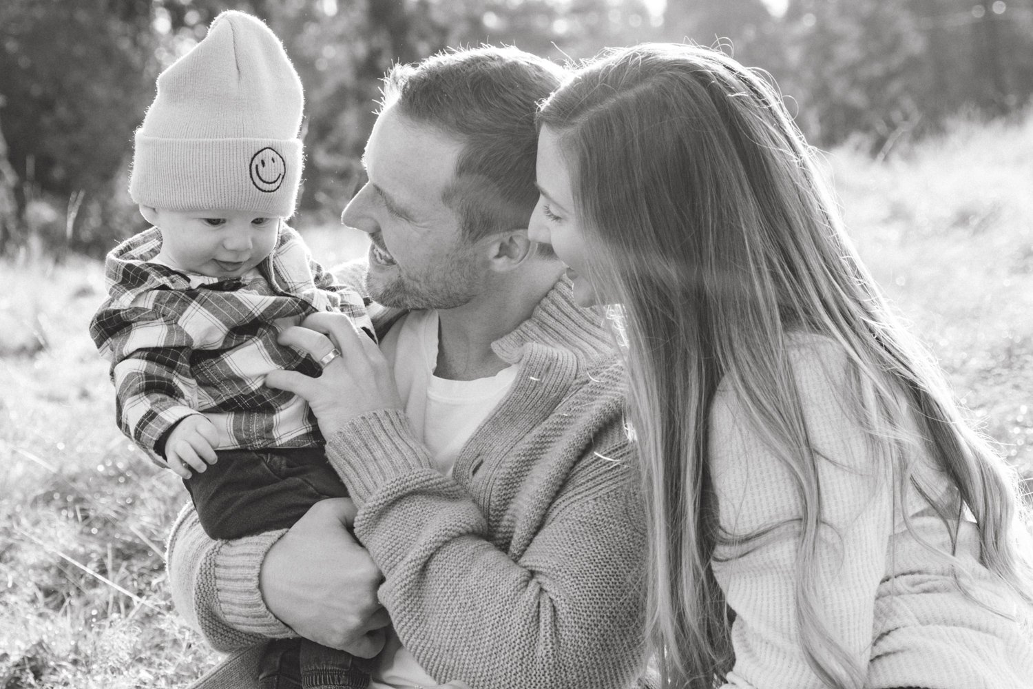 A black and white photo of a happy family outdoors, with a young child in a beanie and plaid shirt sitting on a man's lap, both parents looking at the child lovingly.