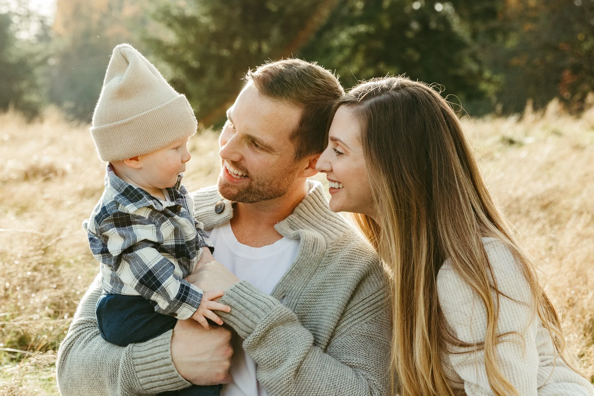 A family of three, a father, mother, and young son, sharing a happy moment outdoors in a grassy field with trees in the background.