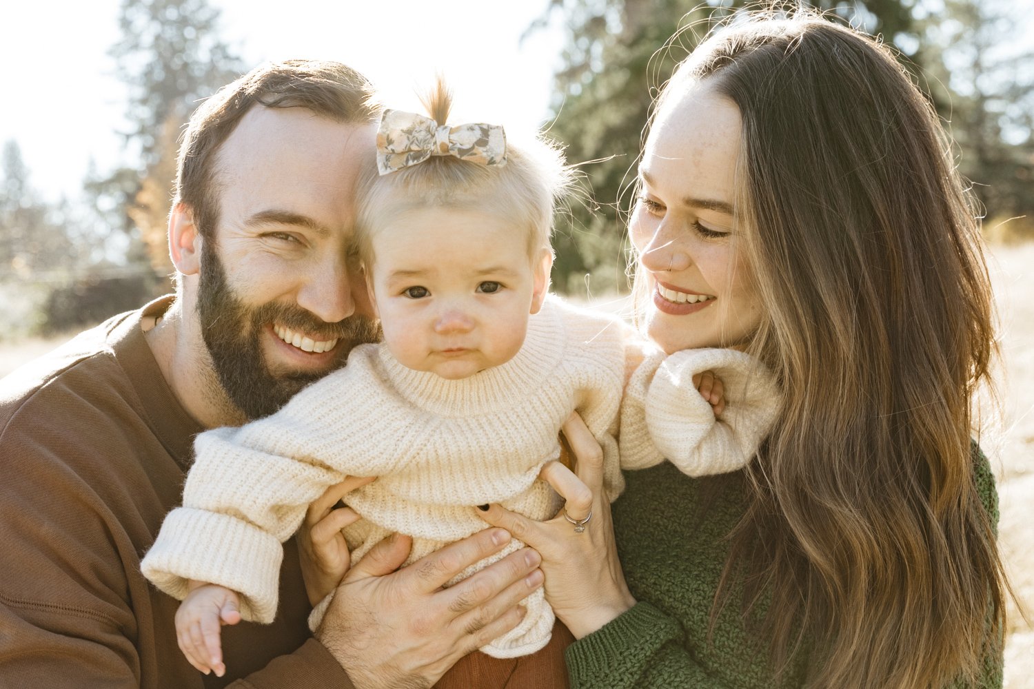 A young family outdoors on a sunny day, with a man, woman, and a toddler girl. The man has a beard and is smiling, holding the toddler. The woman has long brown hair and is smiling at the toddler. The toddler has a neutral expression, wearing a cream