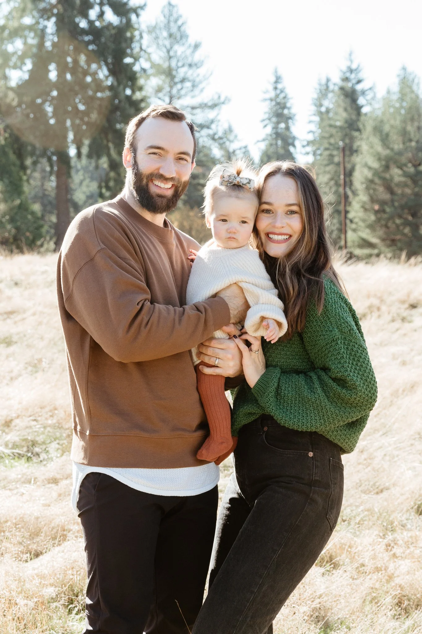 A family of three outdoors in a field of grass and trees, with a man, woman, and young girl posing for a photo, smiling at the camera.