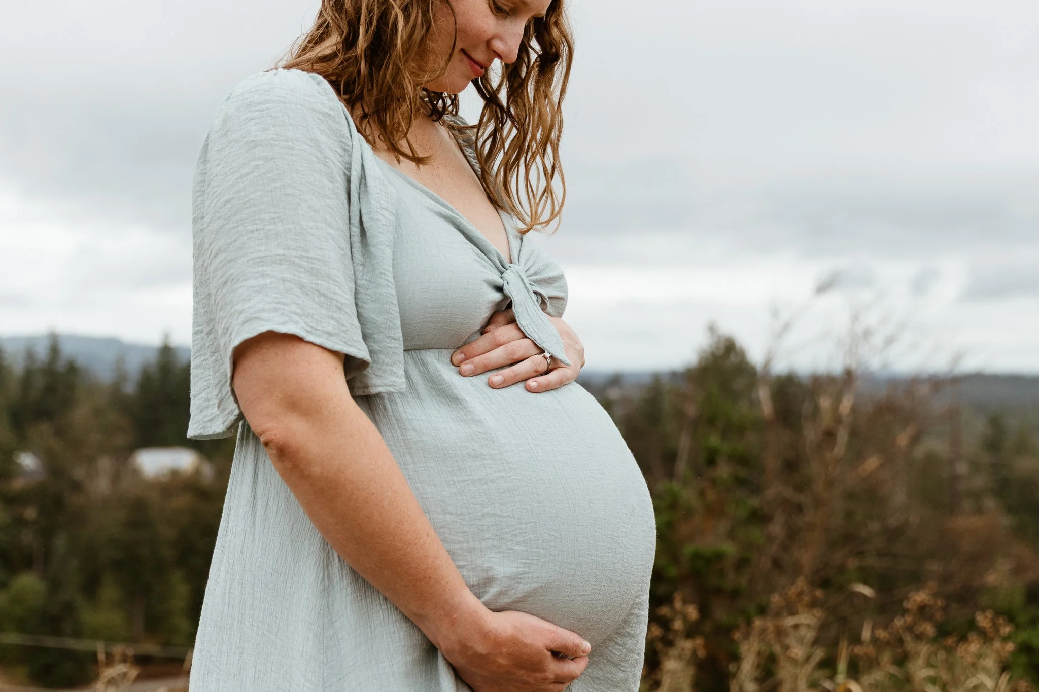 A pregnant woman in a light gray dress, standing outdoors, gently holding her belly with both hands, looking down and smiling softly.