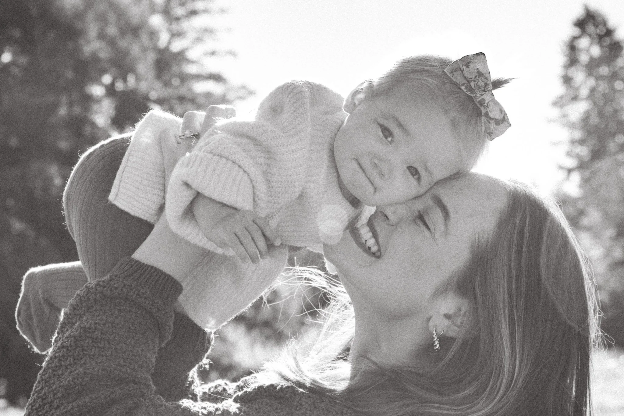 A woman lifting a young girl in the air, smiling and embracing each other outdoors on a sunny day, with trees in the background.