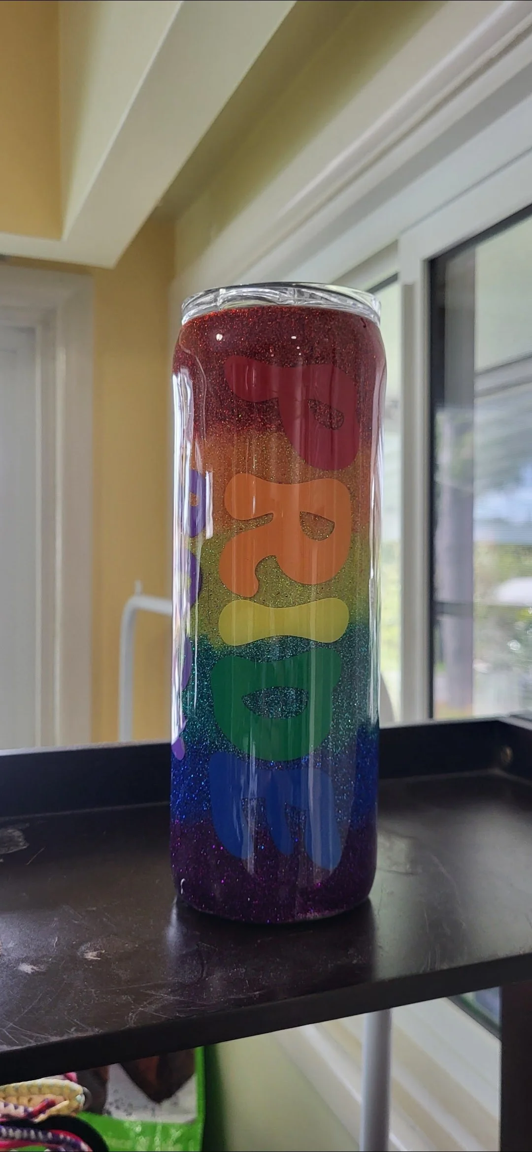 A tall, clear glass with rainbow-colored, glittery shapes inside, placed on a black surface near a window.
