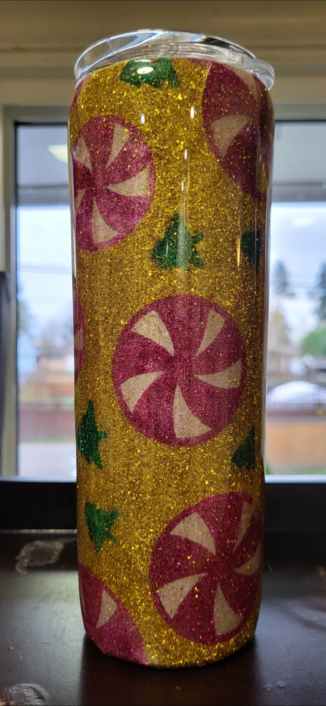 Decorative cylindrical object with red and white peppermint swirl patterns and green holly leaf and berries, covered in gold glitter.
