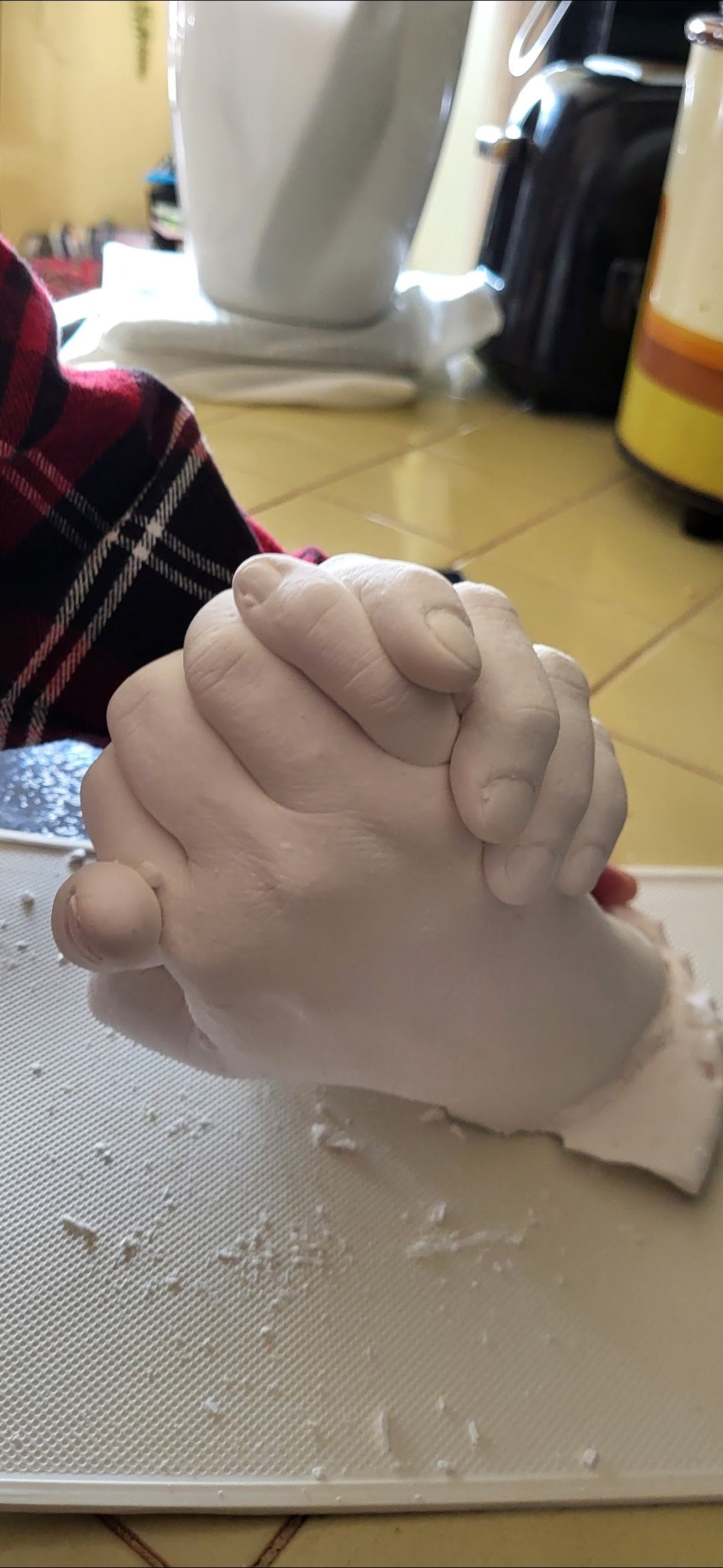 Close-up of two hands clasped together, covered in white dough or similar material, on a kitchen countertop with baking tools in the background.