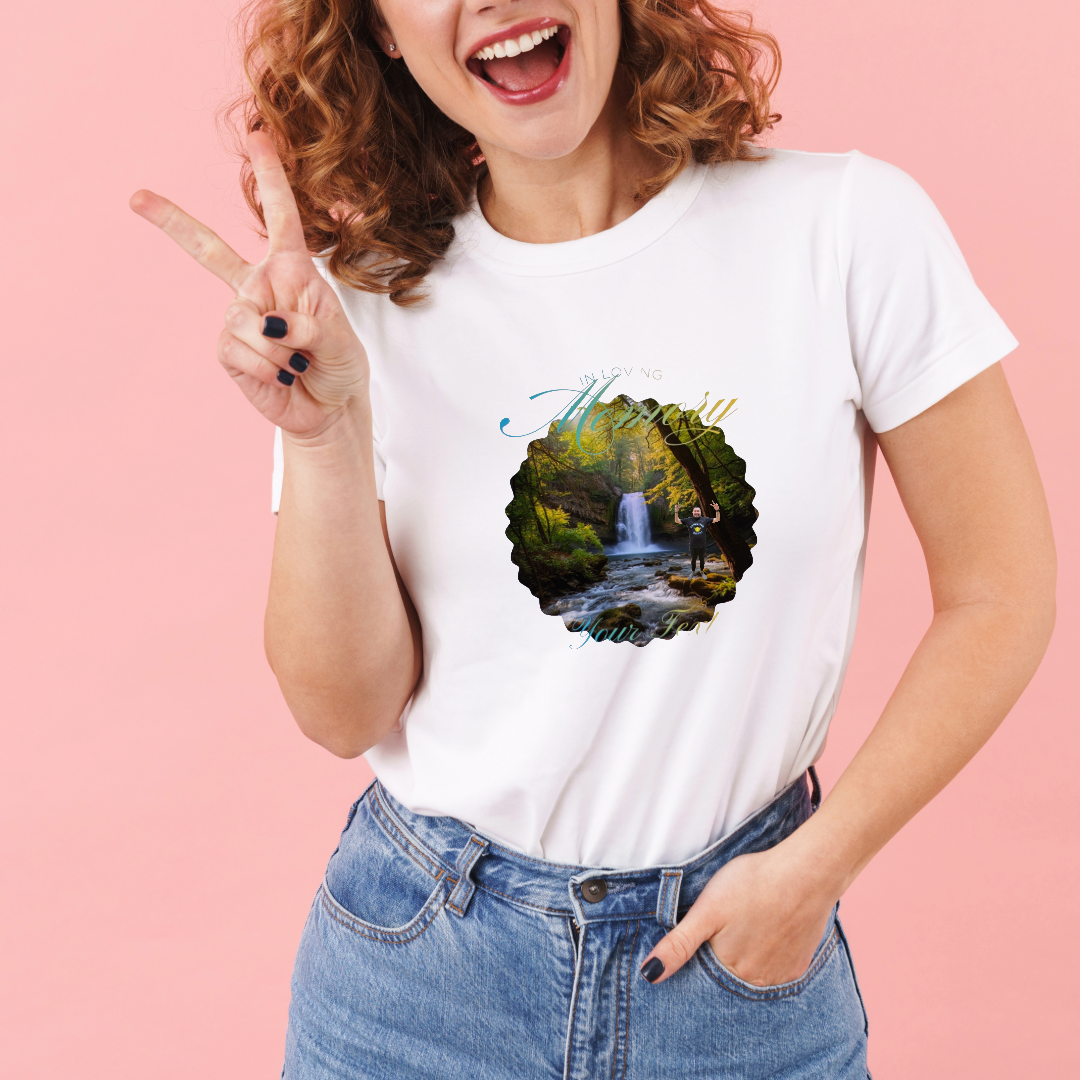Woman smiling and making a peace sign while wearing a white T-shirt with scenic waterfall and forest graphic and placeholder text, against a pink background.