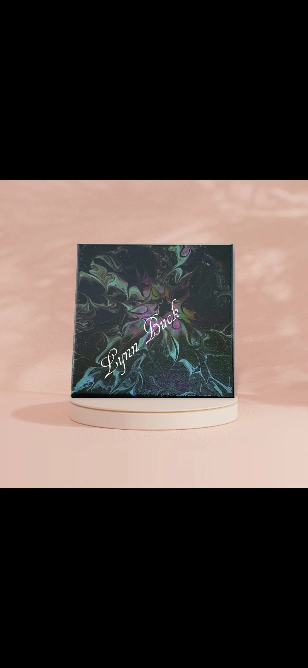A makeup palette with a black, marbled design and the name 'Lynn Buck' written in cursive on the front, placed on a circular white stand against a light pink background.