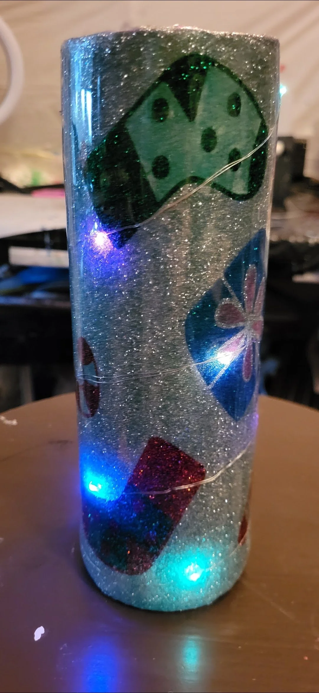 A decorated cylindrical container with glitter, featuring playing card motifs including a Queen of Spades, and colorful LED lights illuminating it.