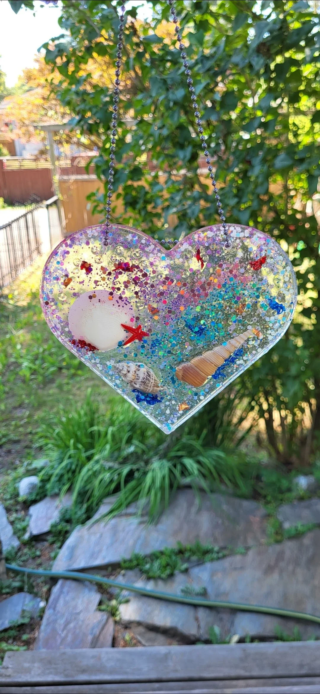 A heart-shaped decorative ornament hanging outdoors, filled with small seashells, a starfish, and glitter.