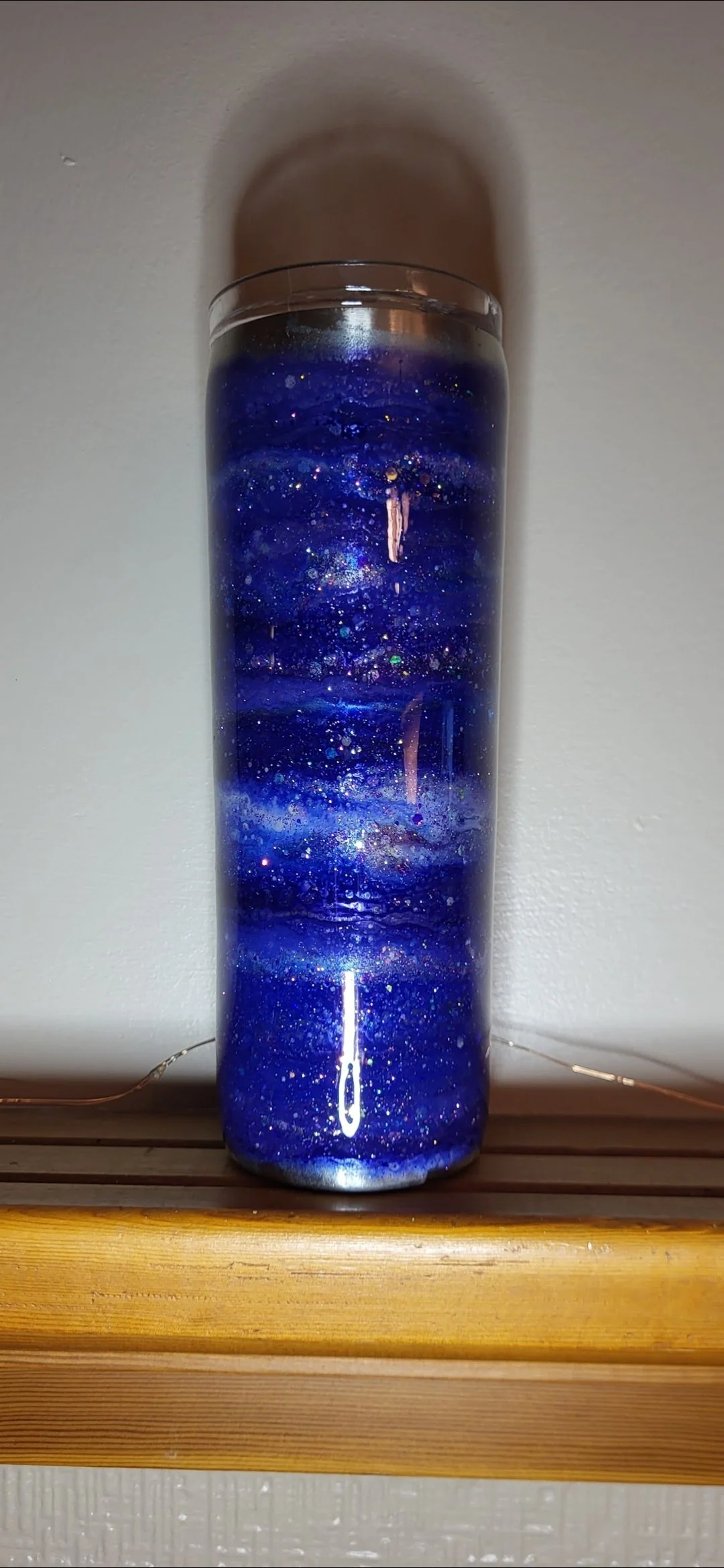 A tall glass filled with a blue galaxy-themed candle or sculpture, featuring specks and swirls resembling stars and cosmic dust, casting a shadow on the wall behind it.