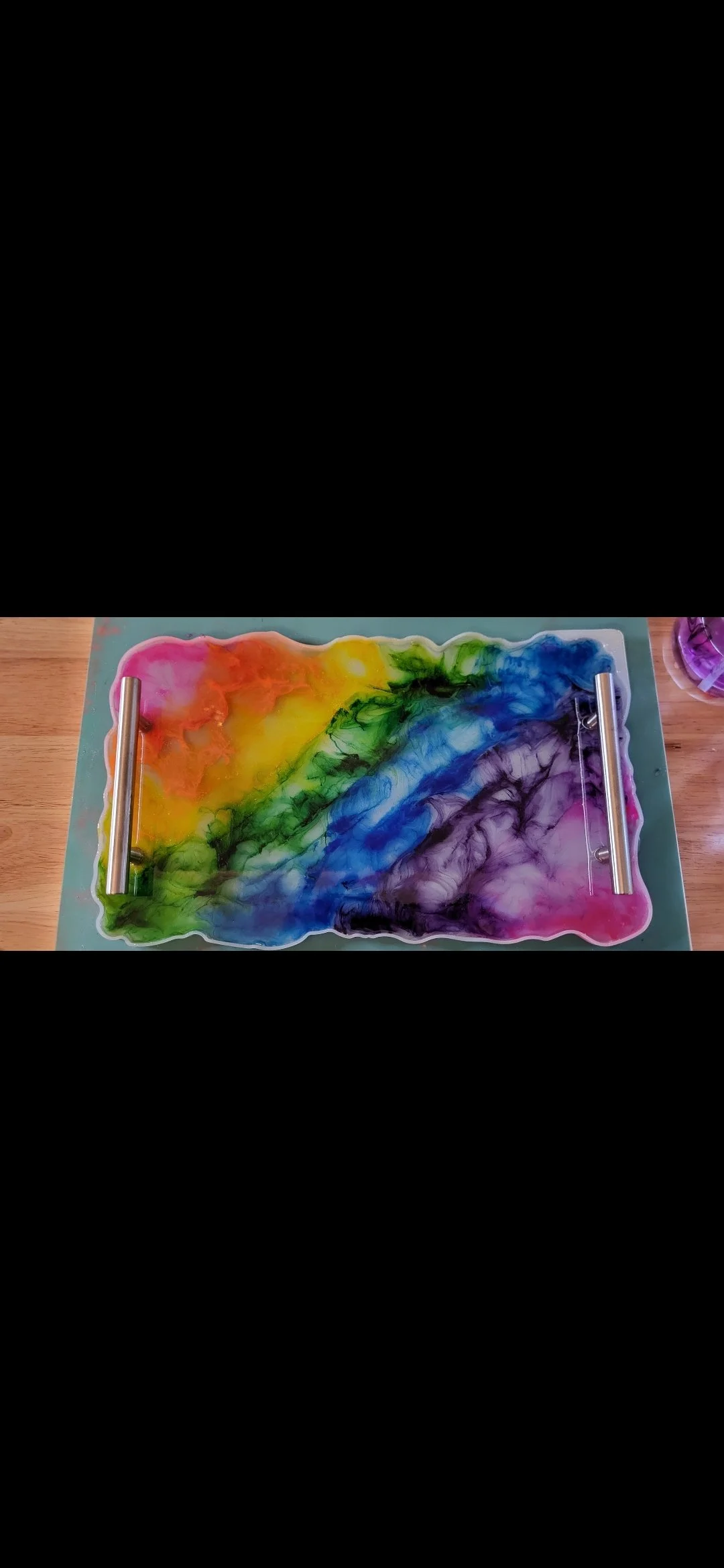 Colorful marble art piece with rainbow colors on a rectangular tray, placed on a wooden surface. Two metallic rollers are positioned on either side of the tray.