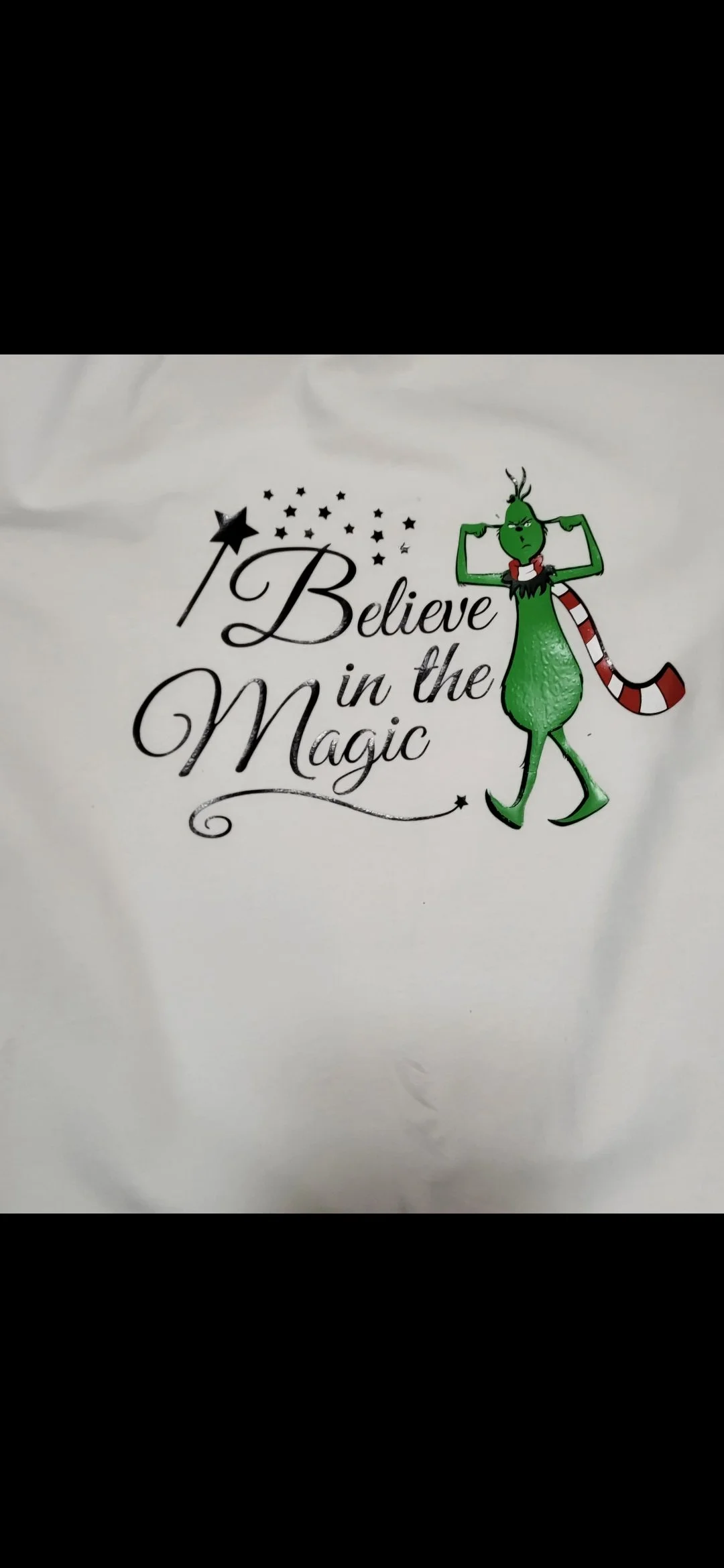 White fabric with a graphic of the Grinch standing with his hands on his head, wearing a Santa hat, red and white striped scarf, and green body. The text says "I Believe in the Magic" with stars around the phrase.
