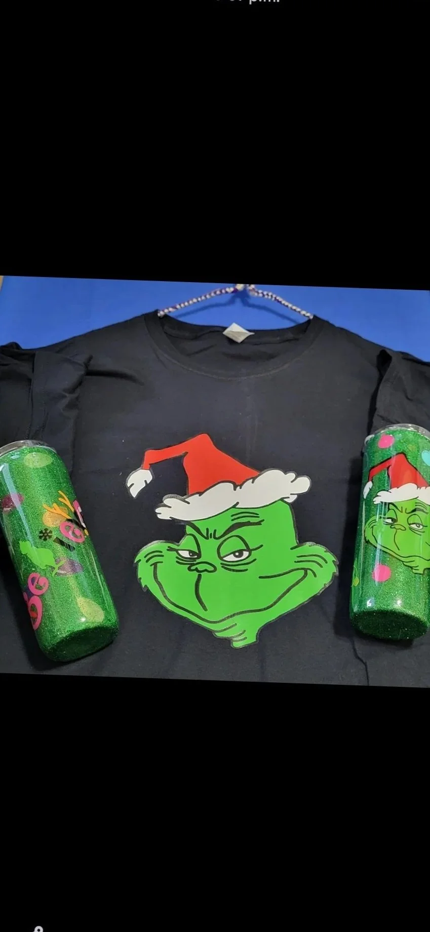 A black T-shirt featuring the Grinch wearing a Santa hat with white fur trim, placed on a blue surface, flanked by two green Coca-Cola cans decorated with pink and green holiday-themed designs.