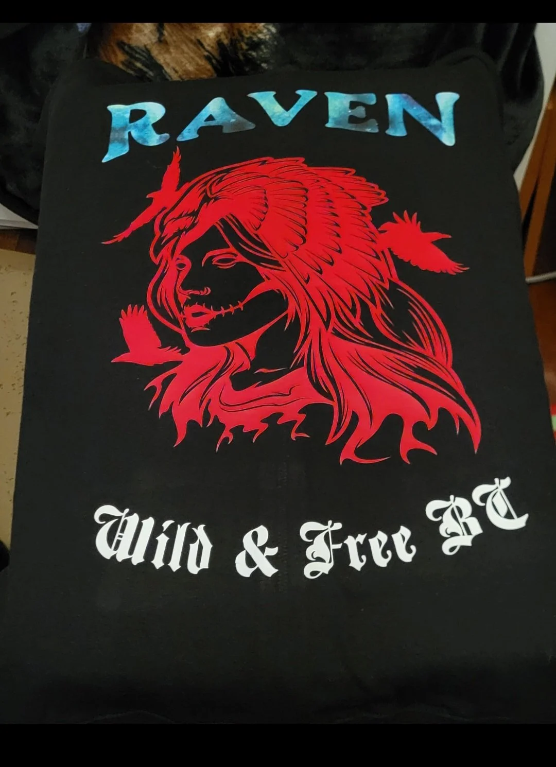 Black T-shirt featuring a red graphic of a woman's face with flowing hair, feathers, and a horn. The shirt has the word 'RAVEN' in blue at the top and the phrase 'Wild & Free TTC' in white Gothic font at the bottom.