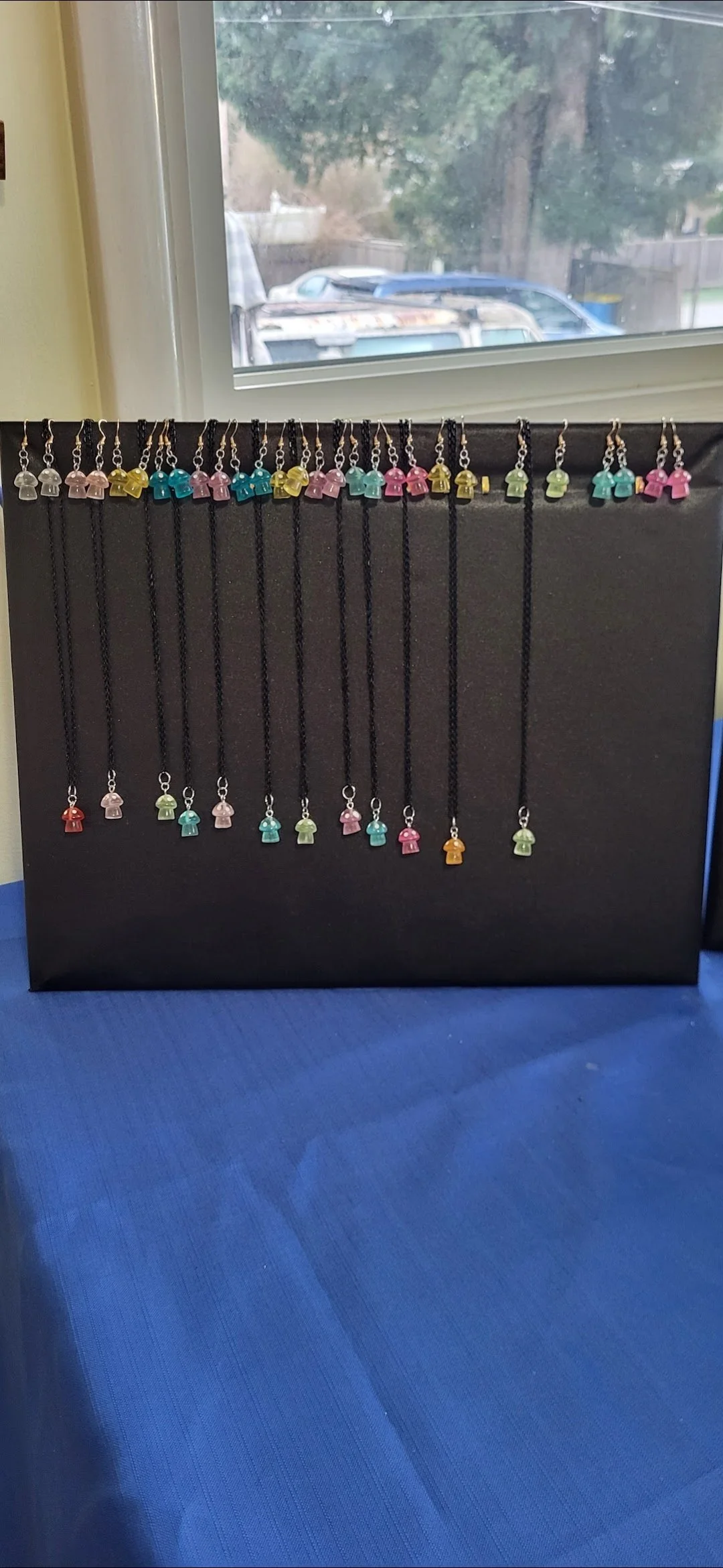 Display of colorful necklace charms shaped like small girls, hanging on a black display board near a window.