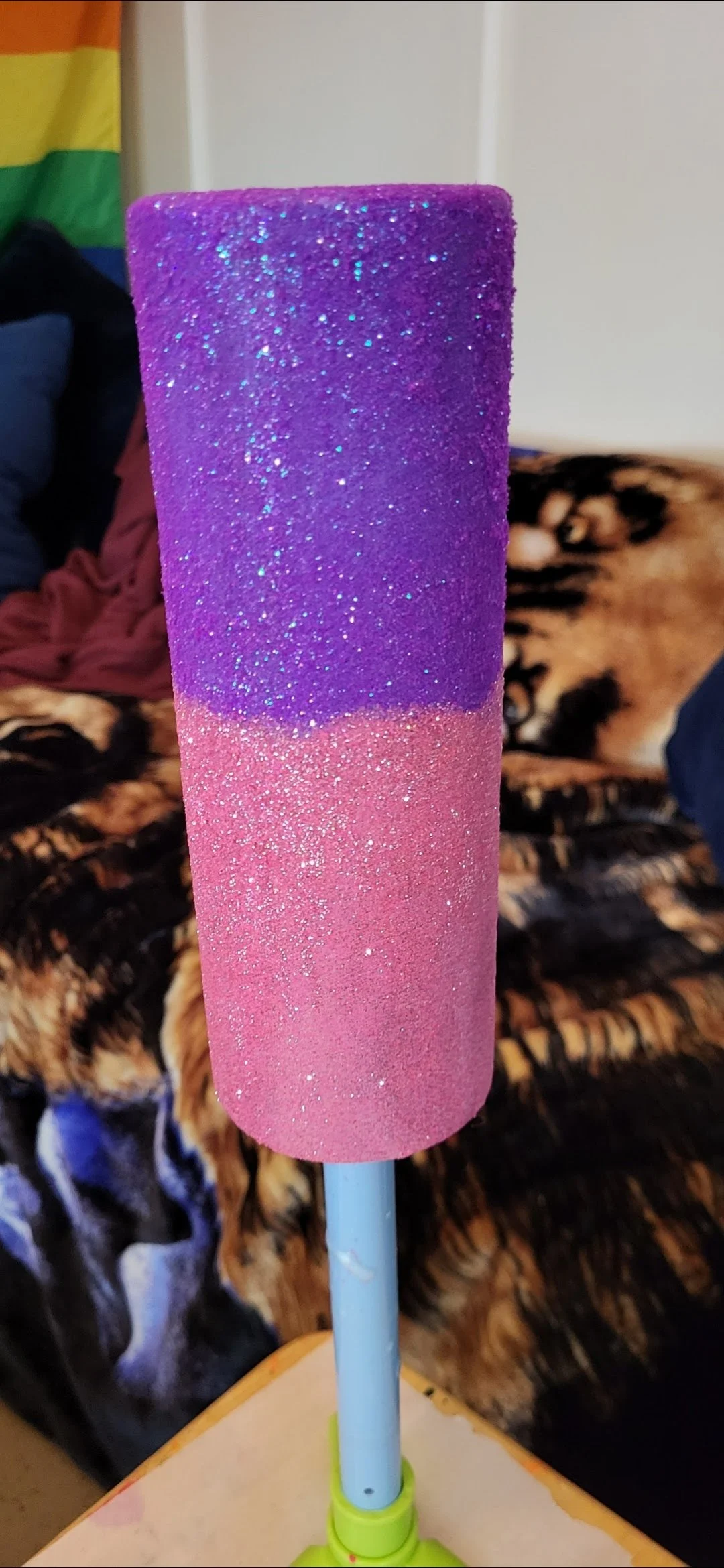 Close-up of a colorful, glittery popsicle with purple and pink sections, mounted on a light blue stick, with a blurred background of a furry blanket and household items.