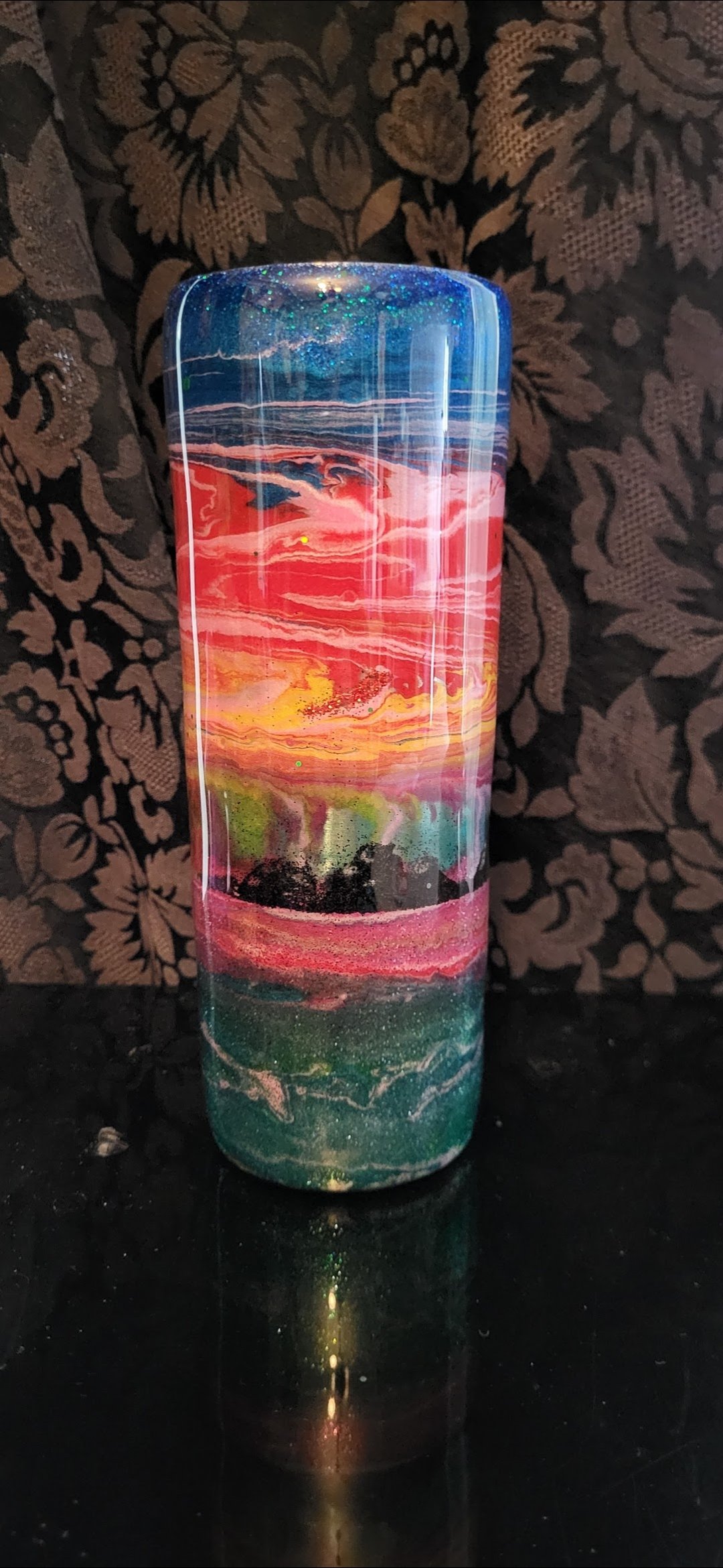 Colorful lava lamp with swirling patterns of blue, pink, yellow, and green against a dark floral patterned background.