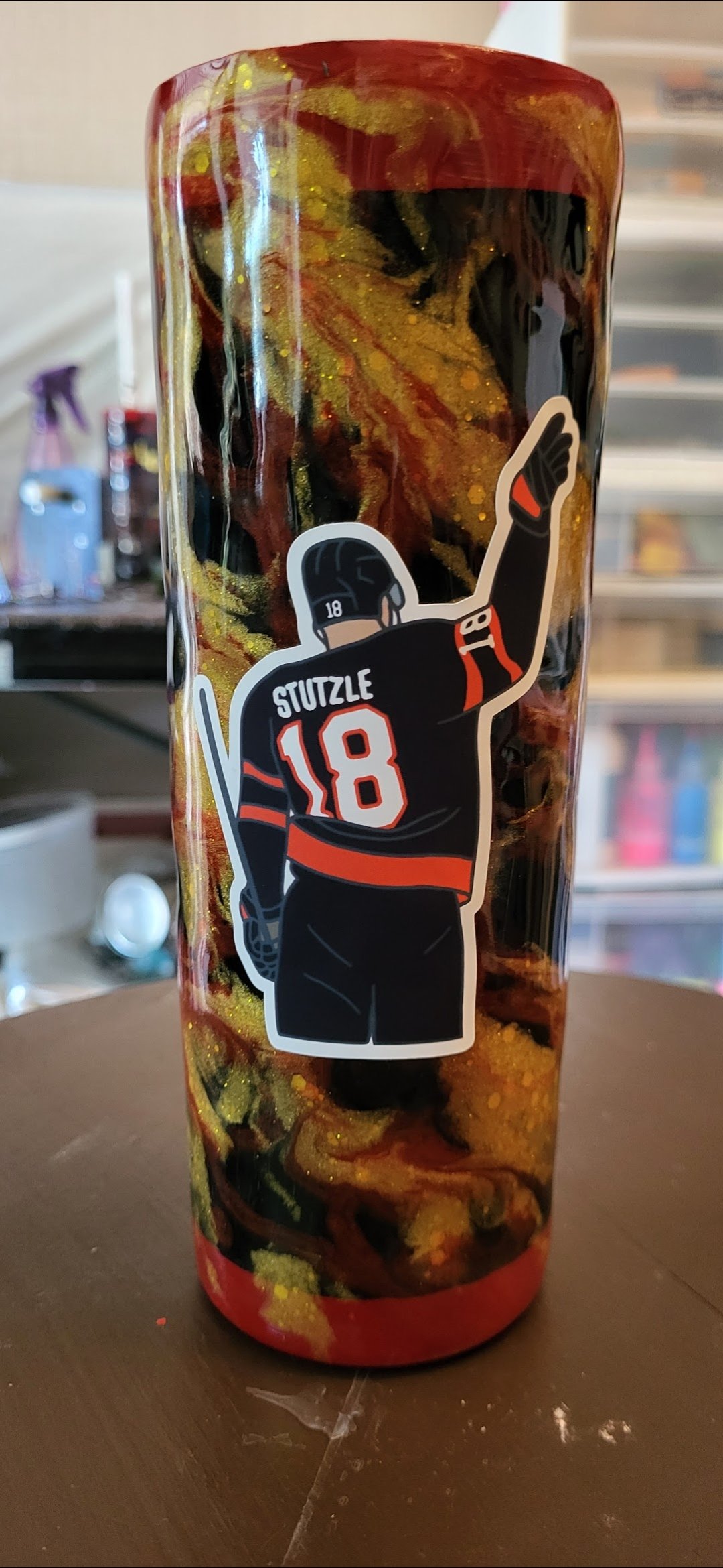 A tall tumbler glass with a black, red, and yellow marbled design, featuring a sticker of a hockey player wearing a black jersey with red accents, number 18, and the name 'Stuzzle,' waving.