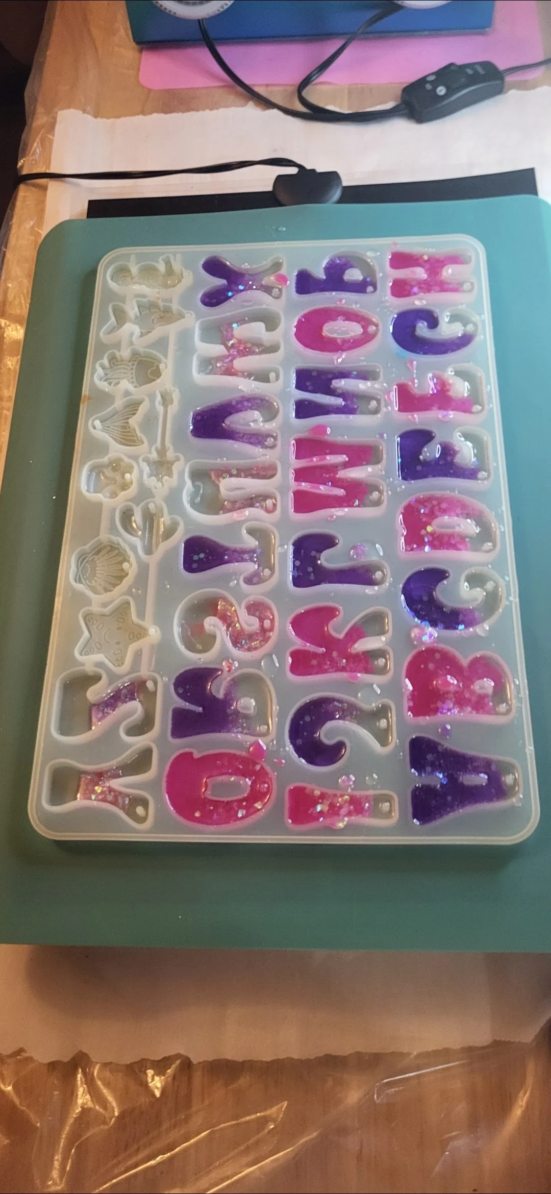 A silicone mold with alphabet letters, filled with pink and purple glittery resin, on a green surface.