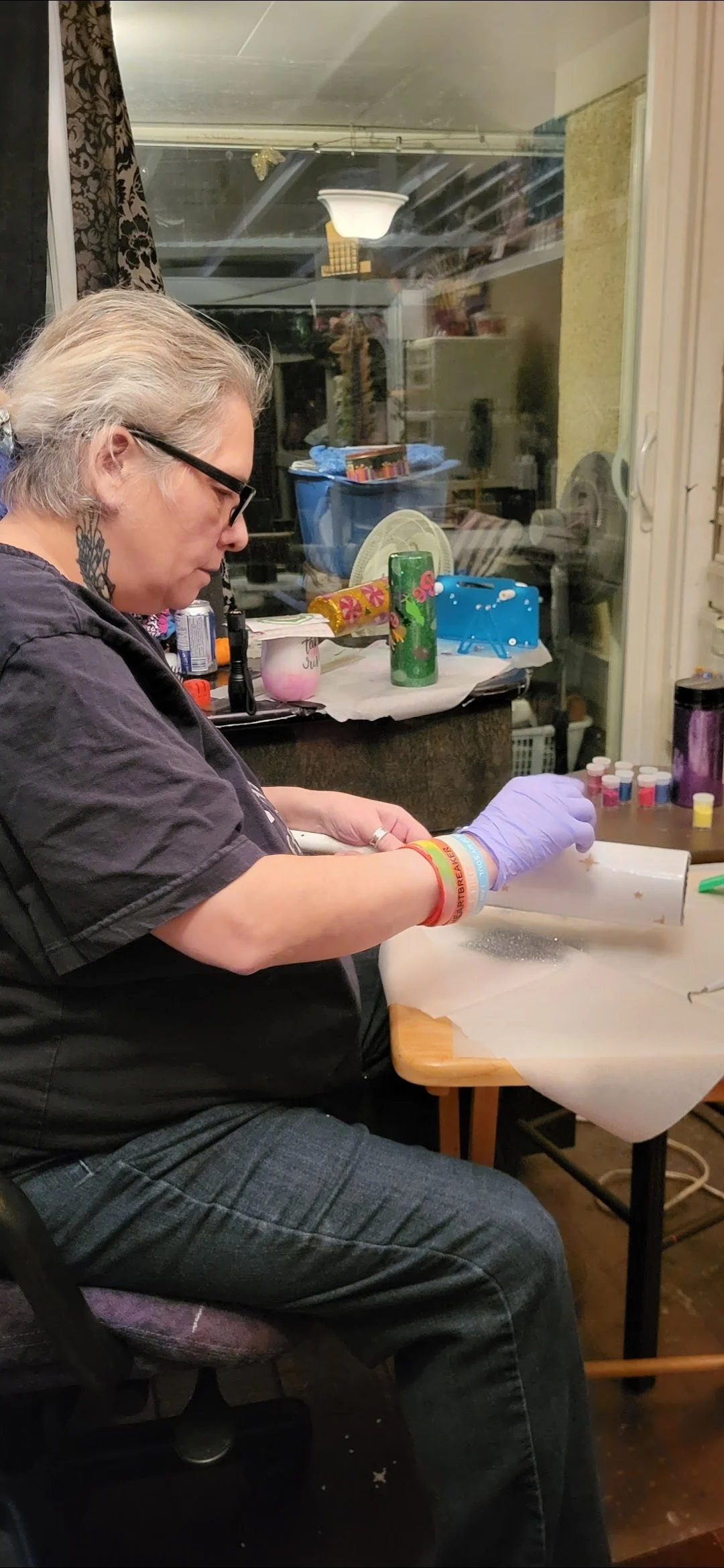 A person wearing glasses, purple gloves, and multiple wristbands is sitting at a table and using a rolling pin to prepare small containers of a pink substance. The background shows a window, a counter with various objects, including a green cylindric