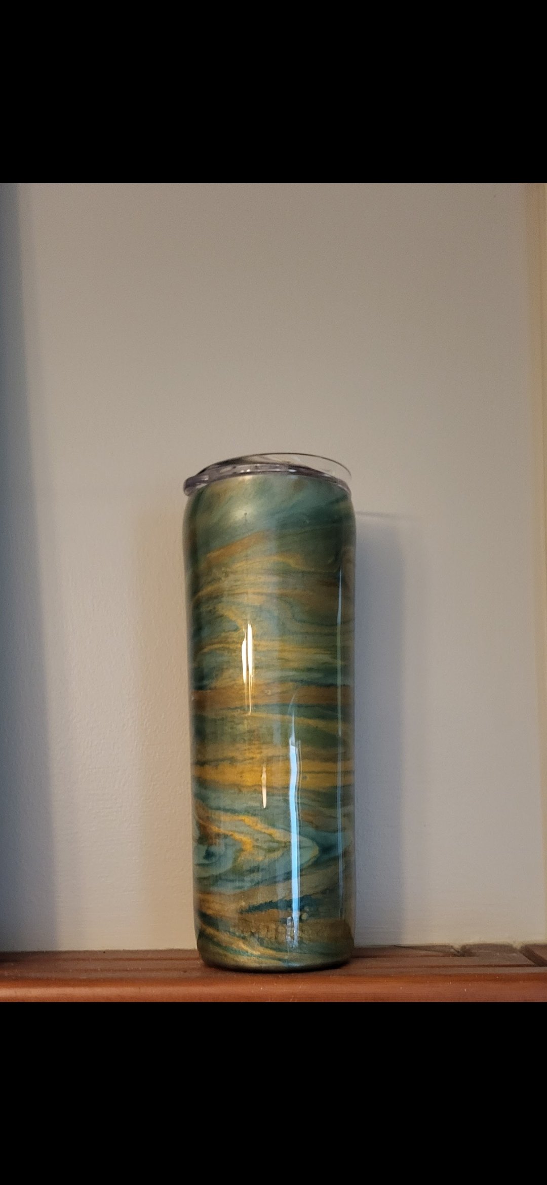 A tall, cylindrical vase with a marbled pattern of blue, green, and yellow hues resting on a wooden surface against a plain, light-colored wall.