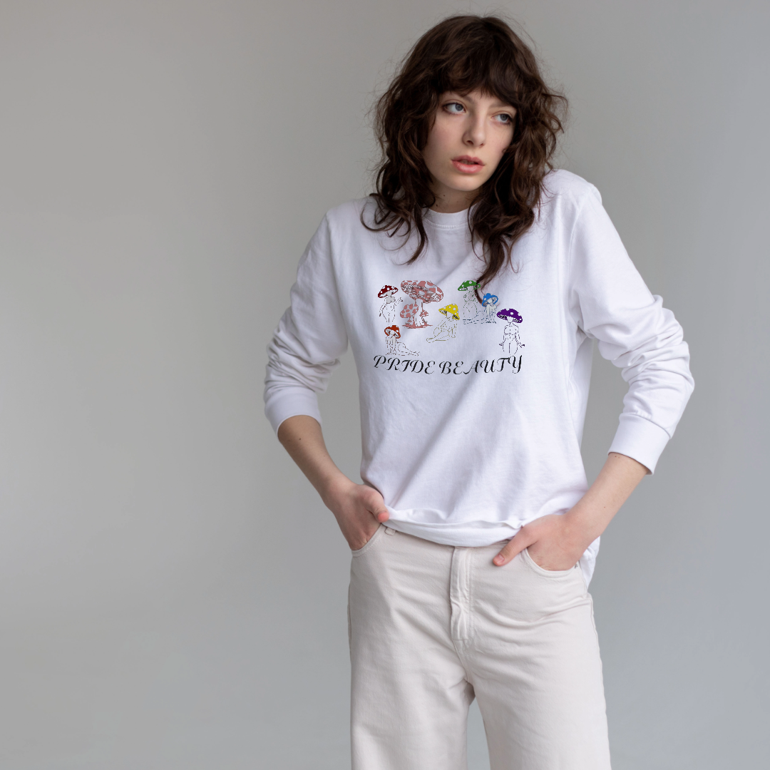 A woman with brown curly hair wearing a white sweatshirt with colorful mushroom illustrations and the phrase 'PRIDE & BEAUTY' in black text.