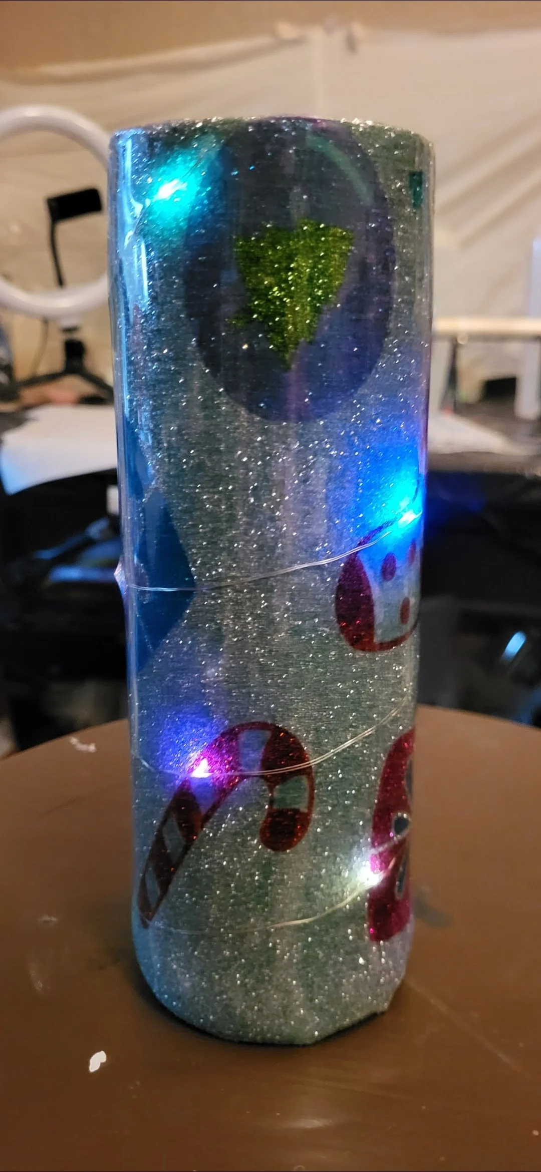 A cylindrical Christmas decoration with embedded shiny glitter, red and green candy cane and holly leaf cutouts, illuminated by blue and purple LED lights.