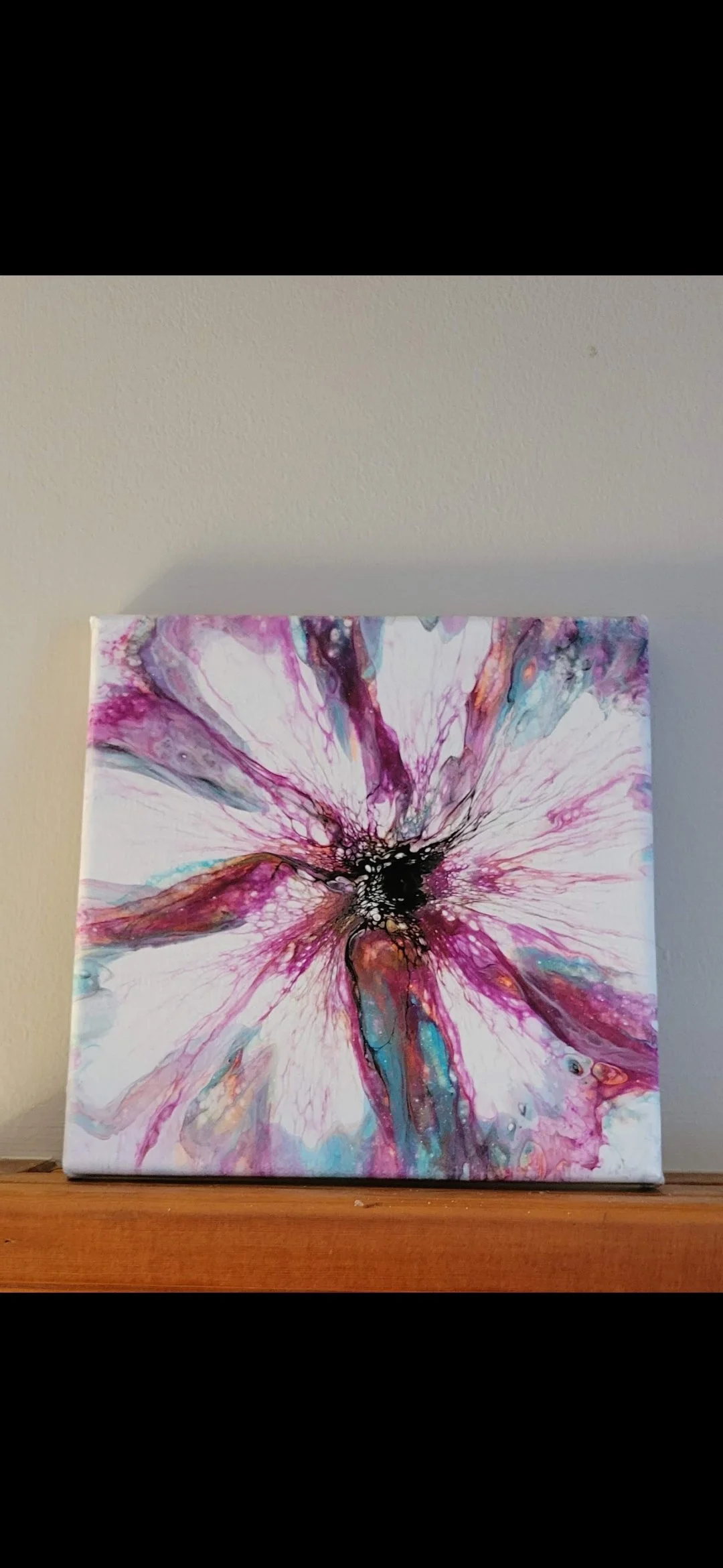 Abstract floral painting with pink, purple, black, teal, and white colors on a canvas, placed on a wooden surface against a light-colored wall.