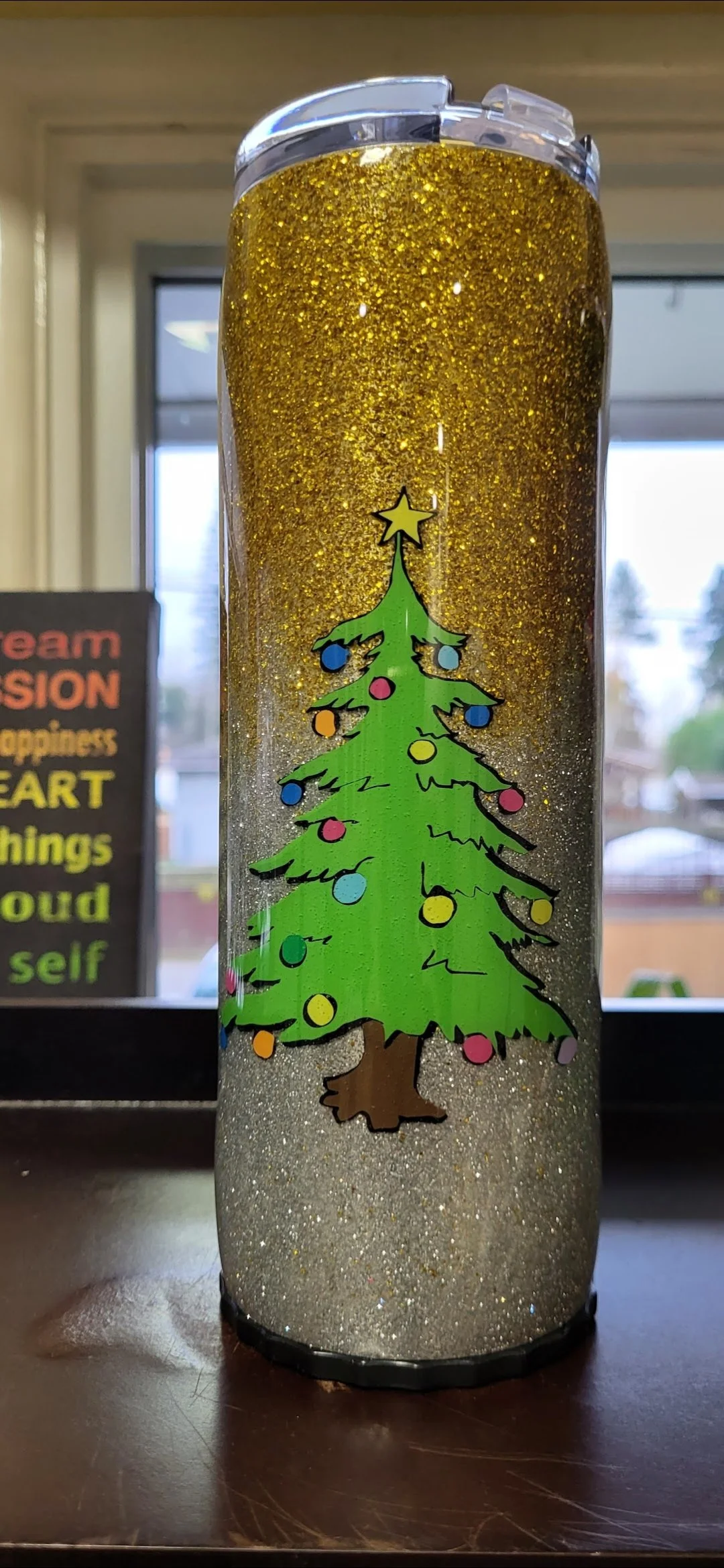A tall tumbler decorated with a Christmas tree design, filled with a glittery gold liquid or substance, with a transparent lid on top.