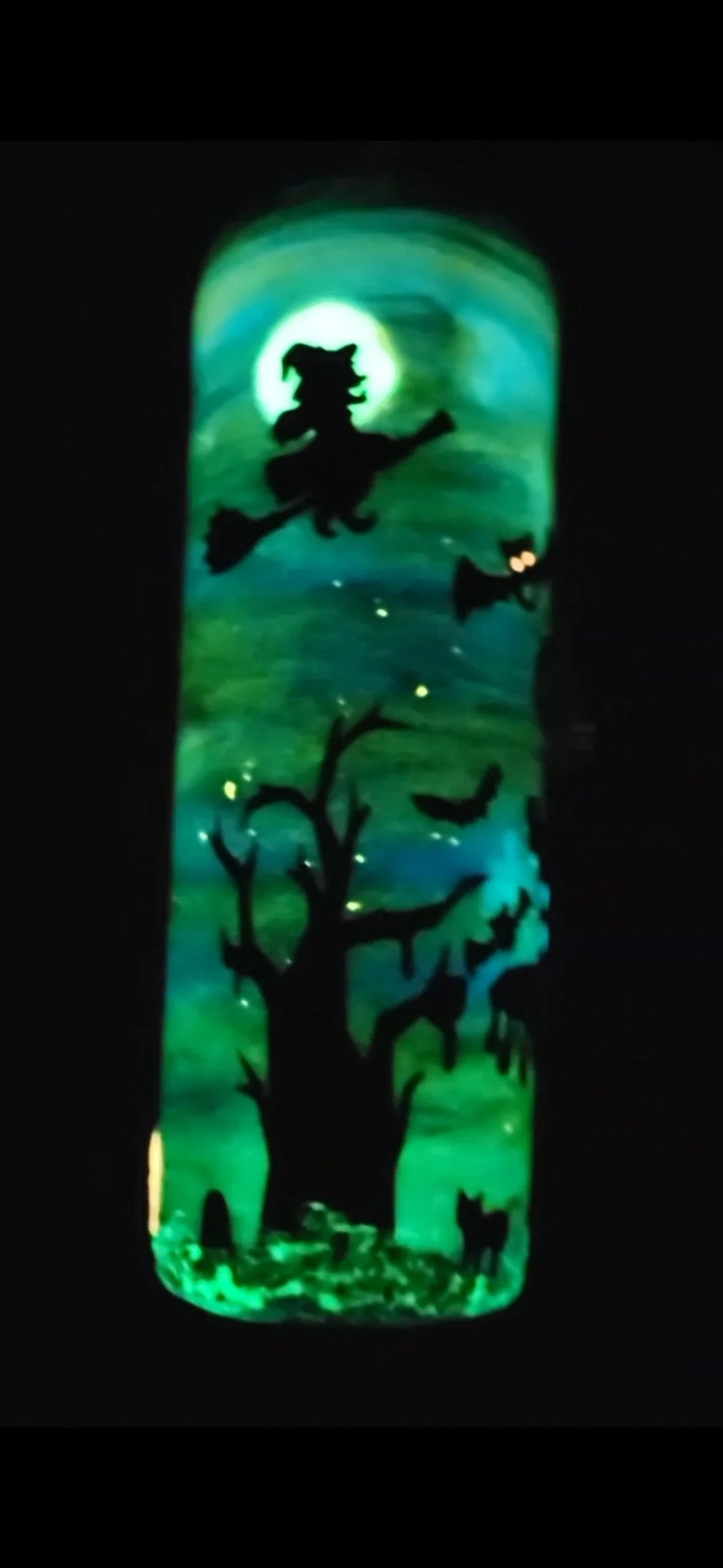 Illuminated stained glass window depicting a witch flying on a broomstick against a moonlit sky with bats and a black cat on a tree.