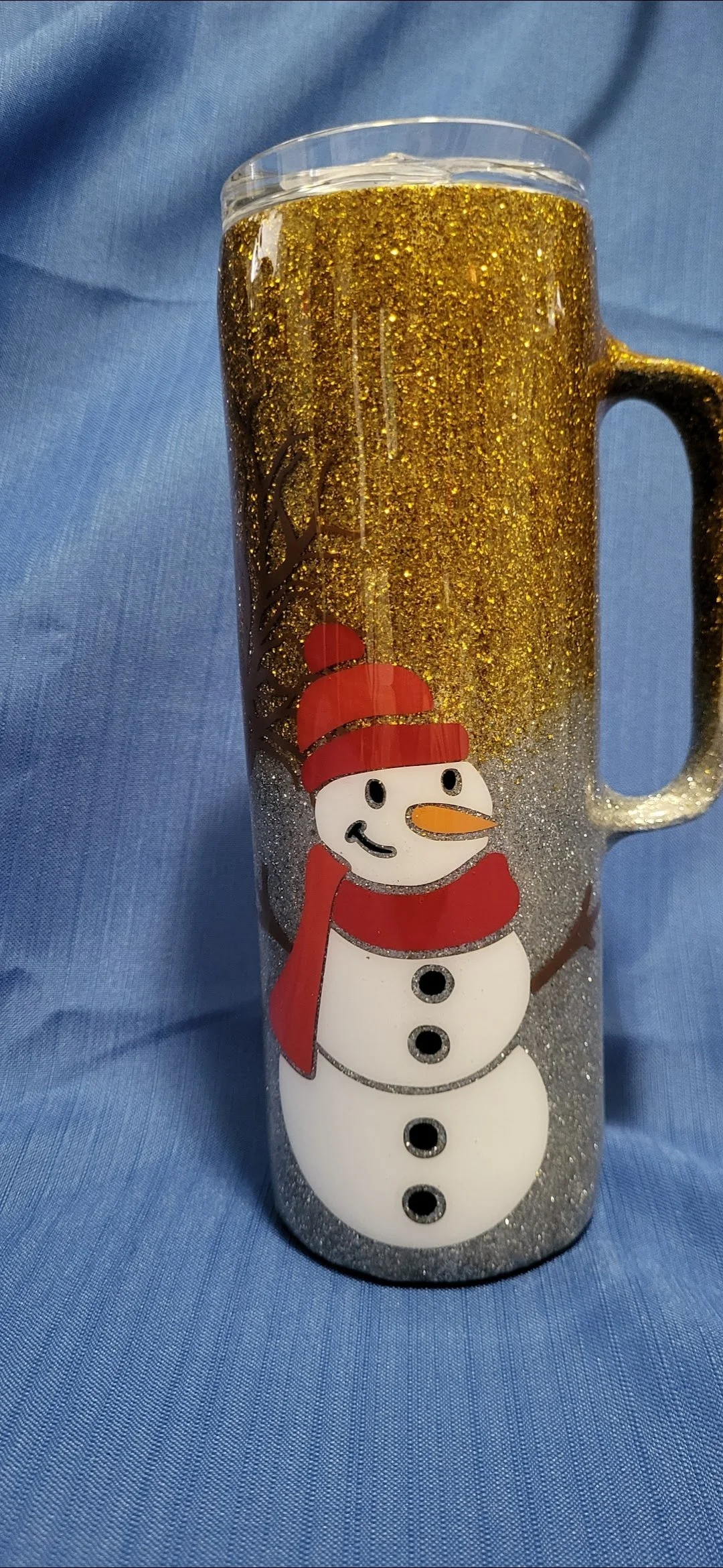 A clear glass tumbler decorated with a snowman wearing a red hat and scarf, filled with a golden glittery liquid, set against a blue fabric background.