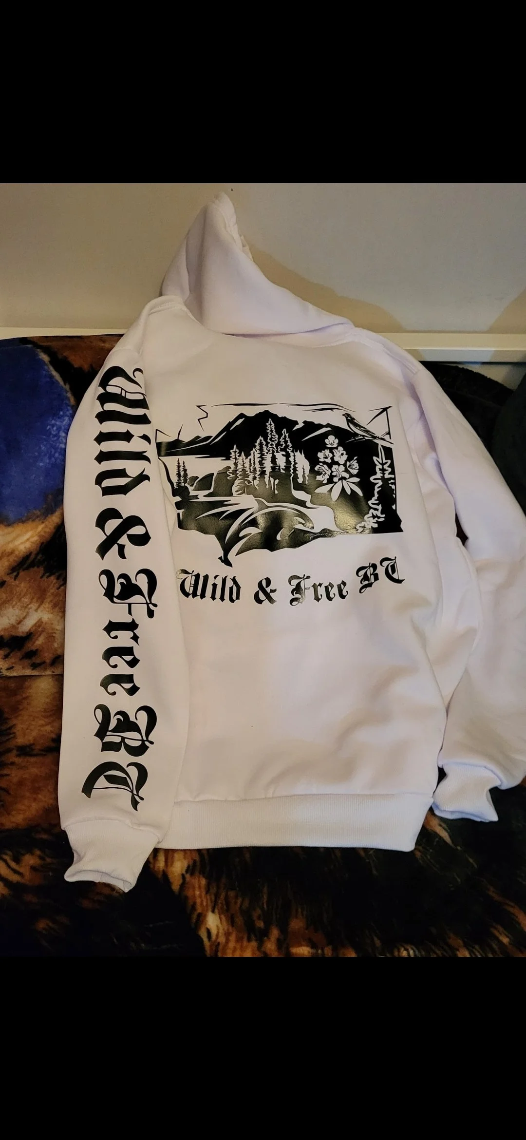 White hoodie with black graphic of mountains, trees, and a river, with text 'Wild & Free BC' on the front and the text 'Deep Roots' on the sleeve.