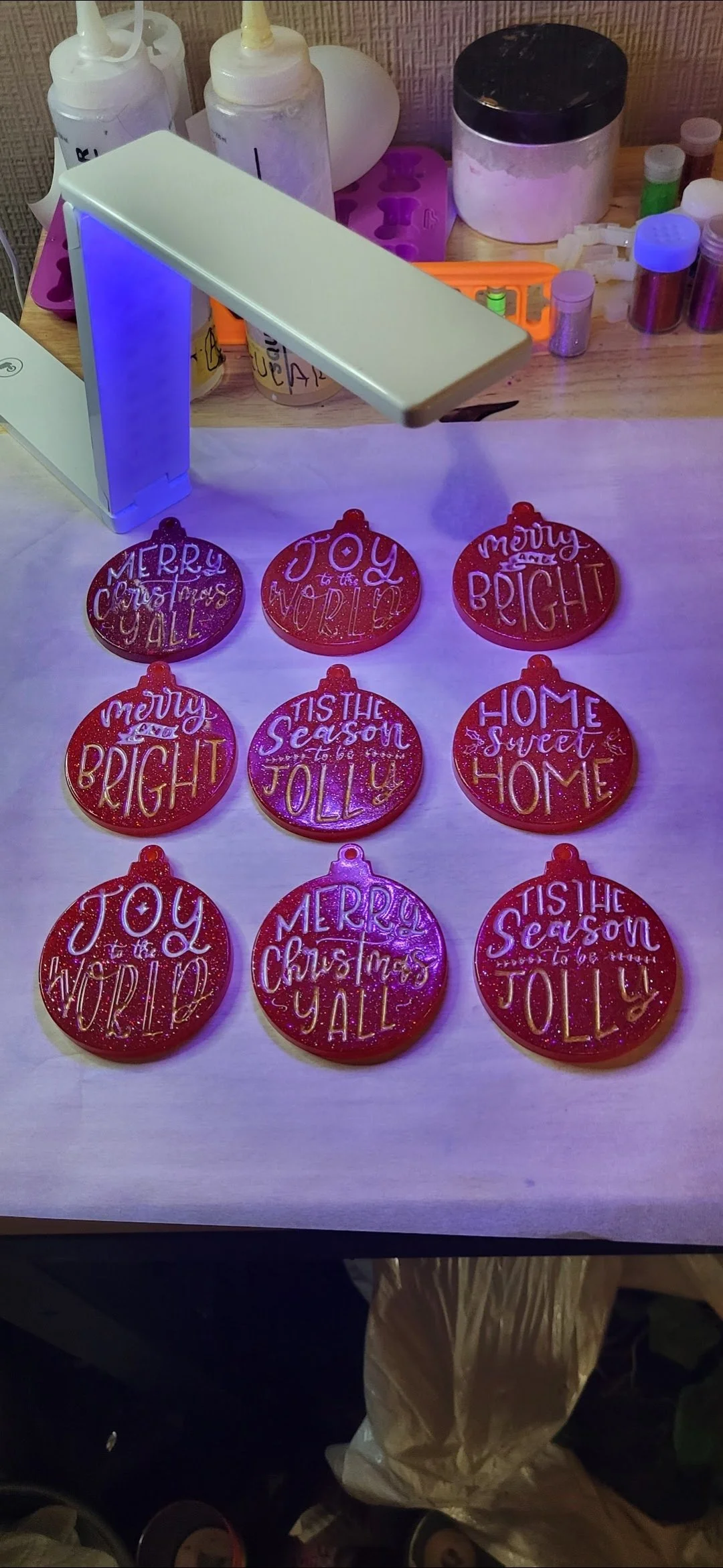 Six red glittery Christmas ornaments with white and purple text on a white surface. The ornaments have holiday greetings like "Merry Christmas Y'all," "Joy to the World," "Tis the Season to be Jolly," and "Home Sweet Home." In the background, there a