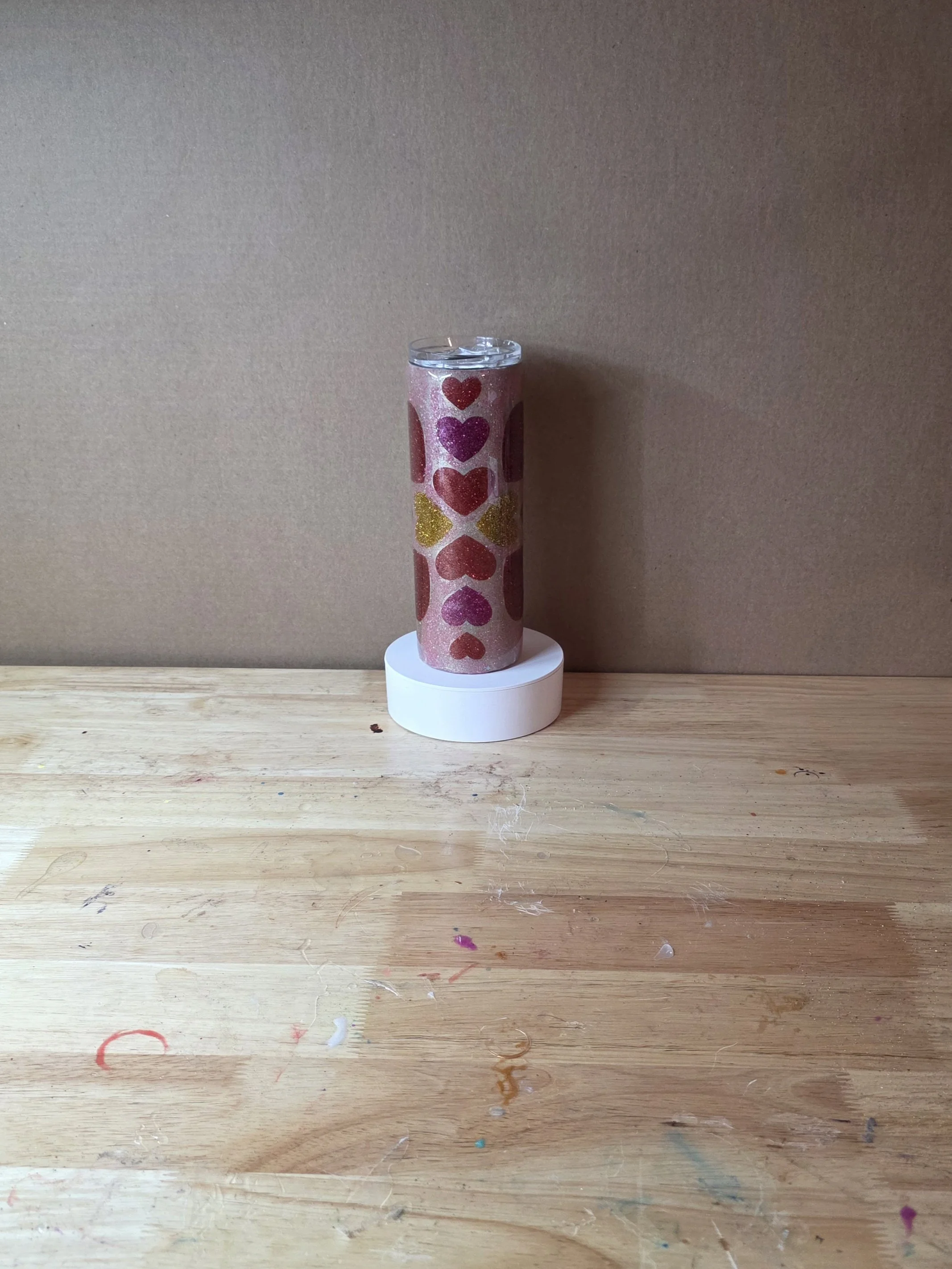 A tall candle with a heart pattern placed on a white circular base on a wooden surface, against a plain beige wall.