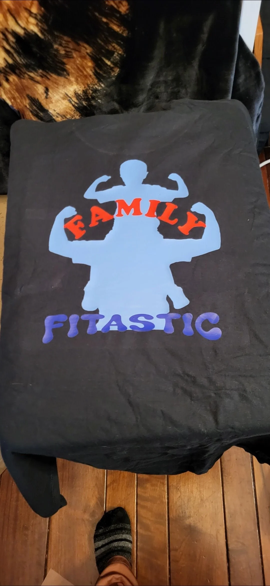 A black T-shirt with a graphic of a person flexing muscles in a classic bodybuilding pose, with the words 'FAMILY FITASTIC' printed on it. The word 'FAMILY' is in red and 'FITASTIC' is in purple, with the letters arranged around the image.